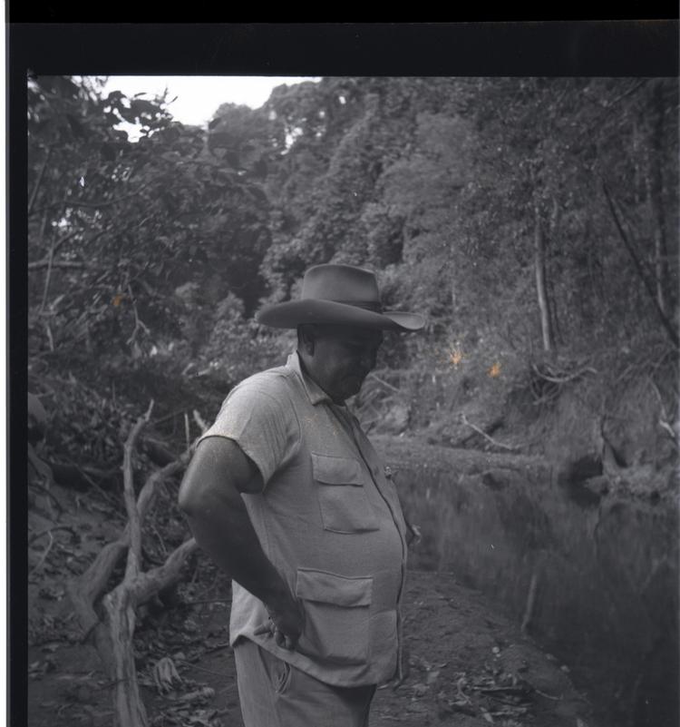 Image of Black and white medium format negative of portly man in hat standing beside a river with trees