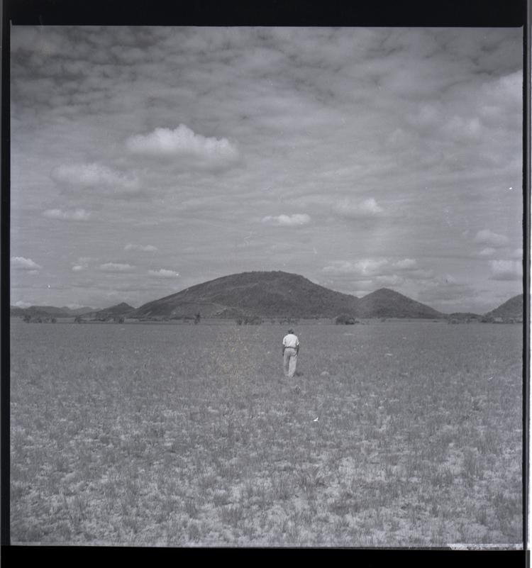 image of Black and white medium format negative of man walking away from camera in flat landscape with mountains in background