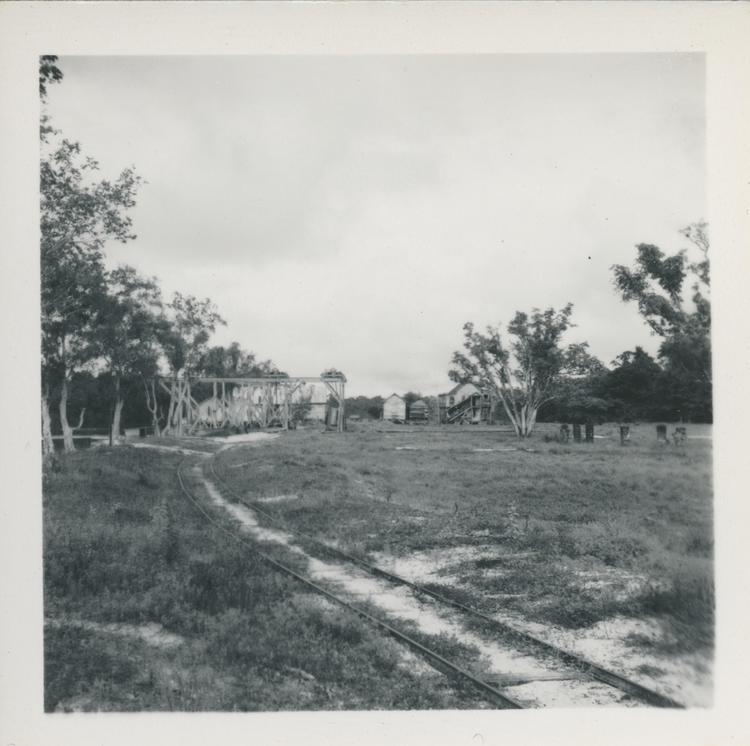image of Black and white print of overgrown metal tracks leading to wooden loading structure and houses by river - a small settlement