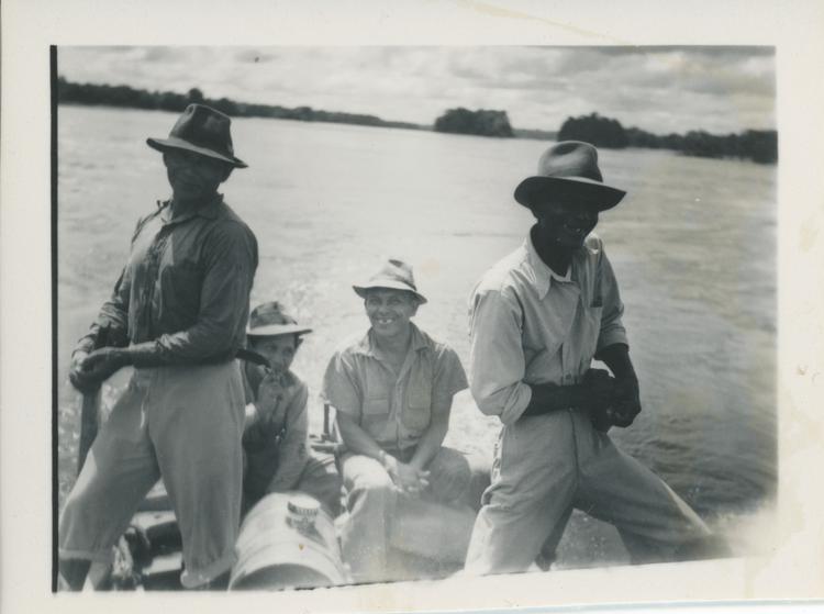 Black and white print of four men in a boat (with motor) with river behind