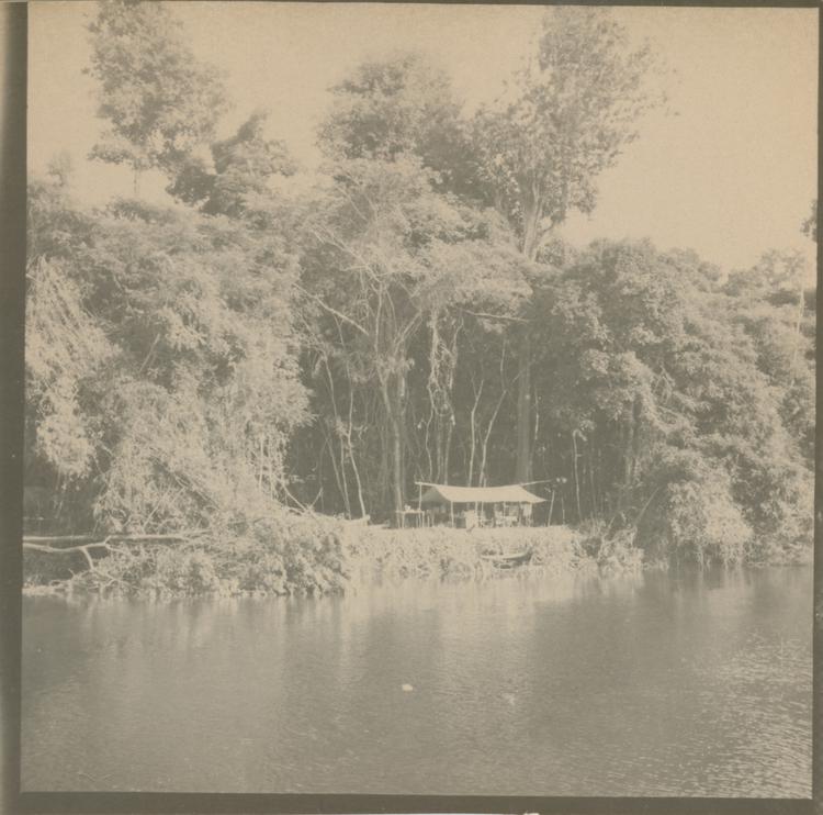 image of Black and white (sepia tones) print of a hut beside a river in a small clearing in tall trees