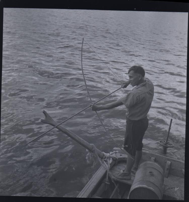 Black and white medium format negative of man standing in a boat aiming a bow and  arrow at the water