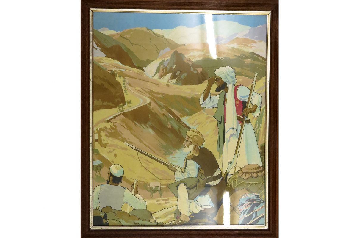 Framed, polychrome print of the Khyber Pass. Pathans with rifles watching travellers on camel-train on the road through the mountains.