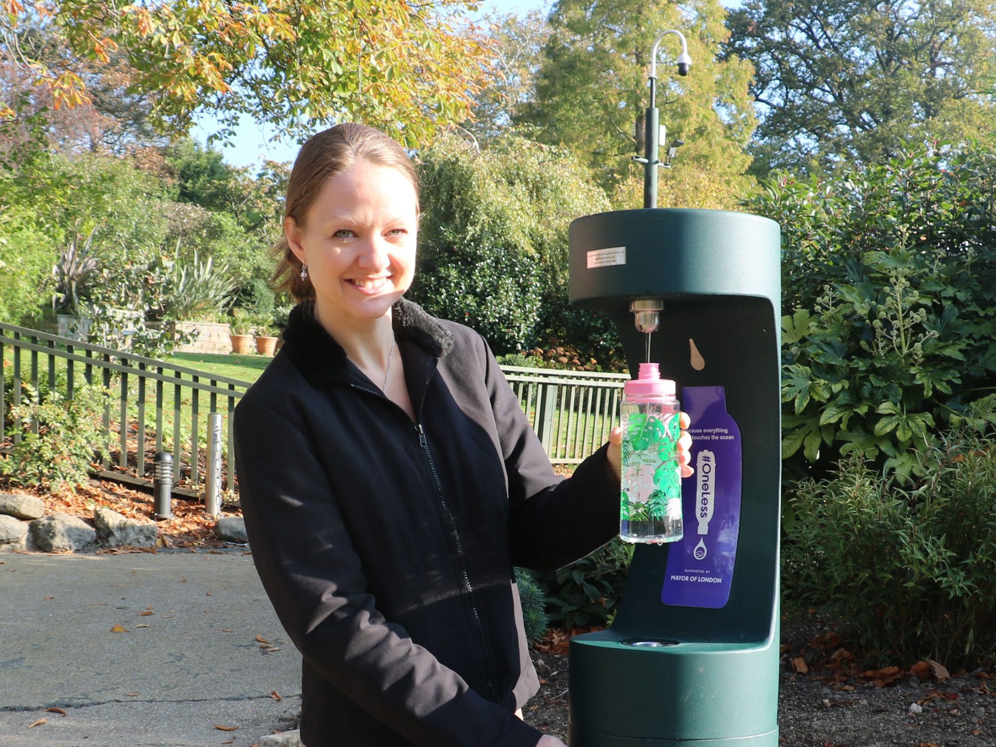 A woman is filling a water bottle at a public drinking fountain and is smiling at the camera. She is in a Garden.
