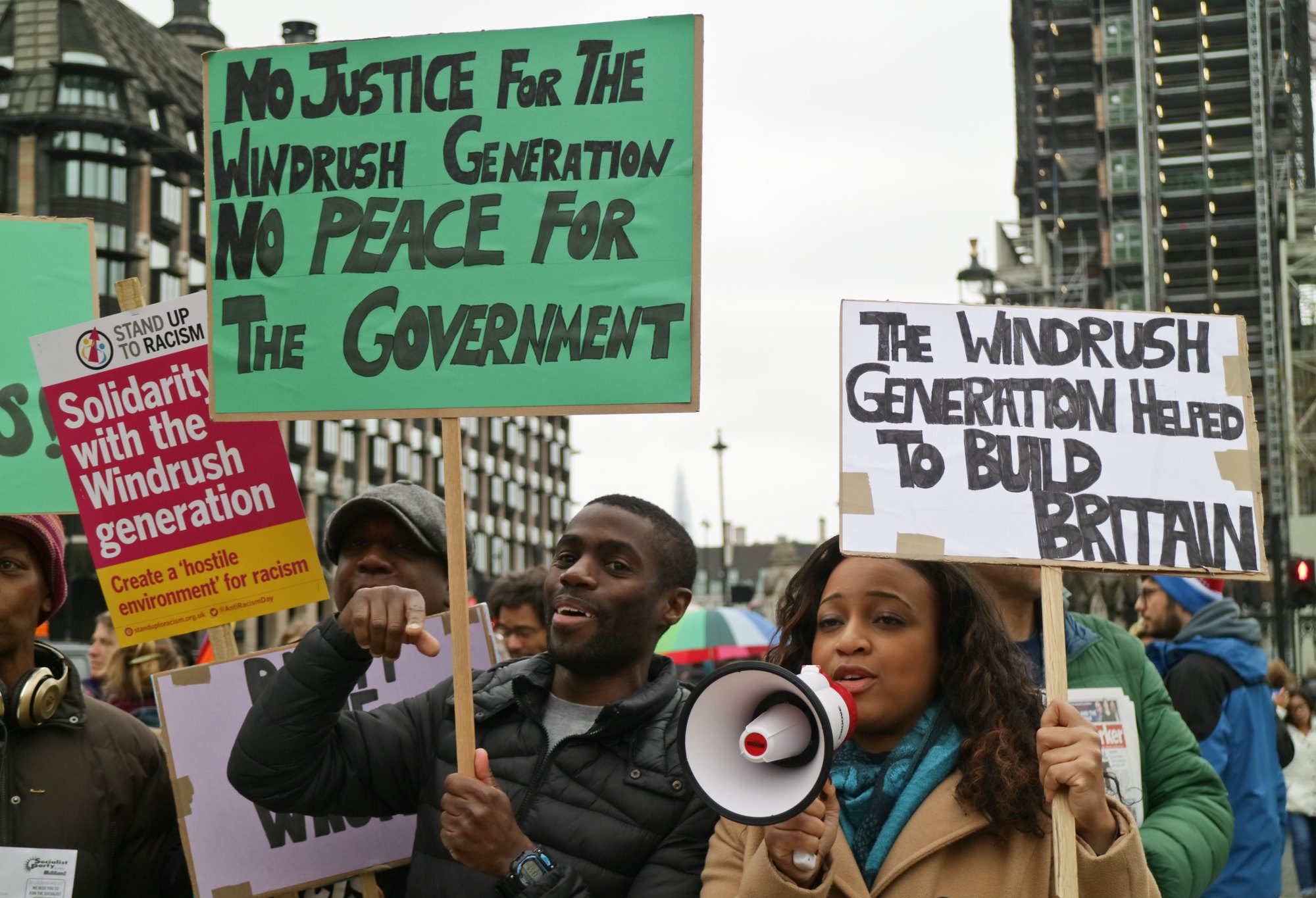 A group of protesters in London. Towards the camera is a woman and a man, both holding placards. The woman is talking into a loudhailer.