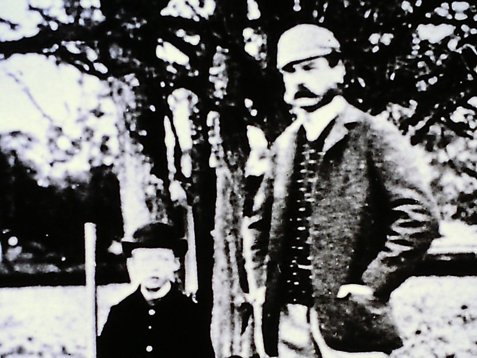 A black and white picture of a man with a gun standing next to a boy who is holding a gun. The man is wearing smart clothes and has a hat and moustache