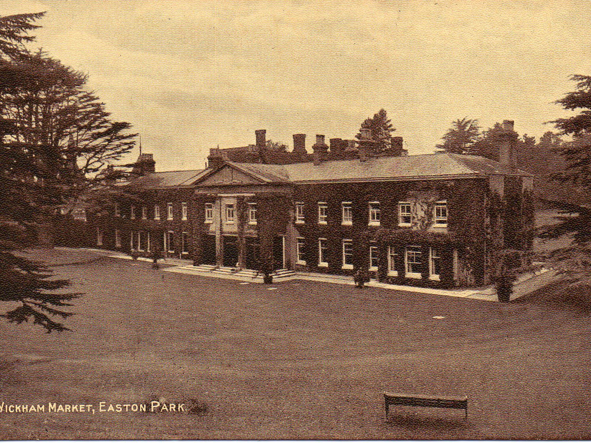A sepia postcard showing a long two story grand house covered in ivy with a lawn surrounding it and trees