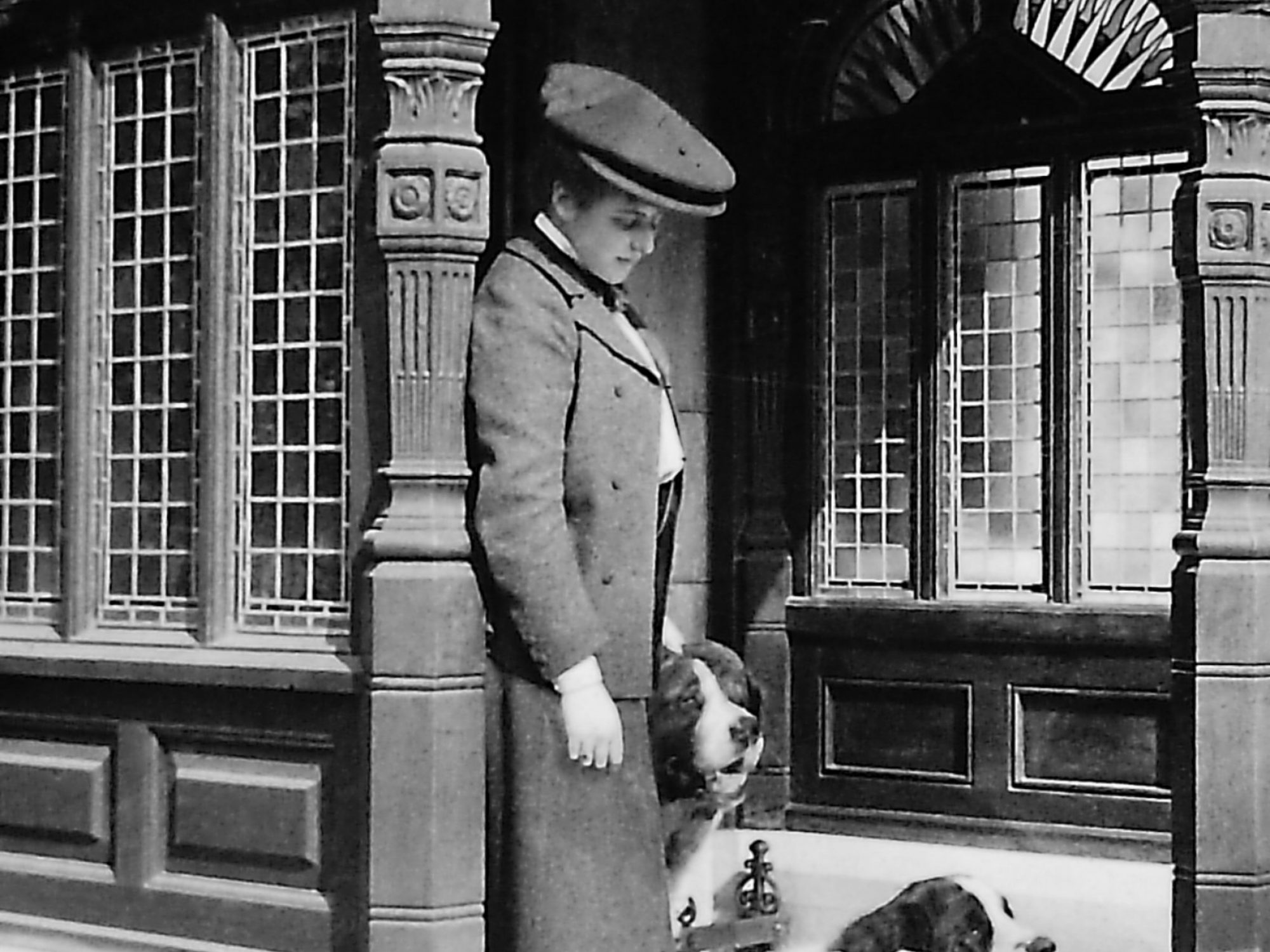 A black and white shot of a woman stood in a doorway in edwardian clothes. She has three dogs at her feet which she is looking down at
