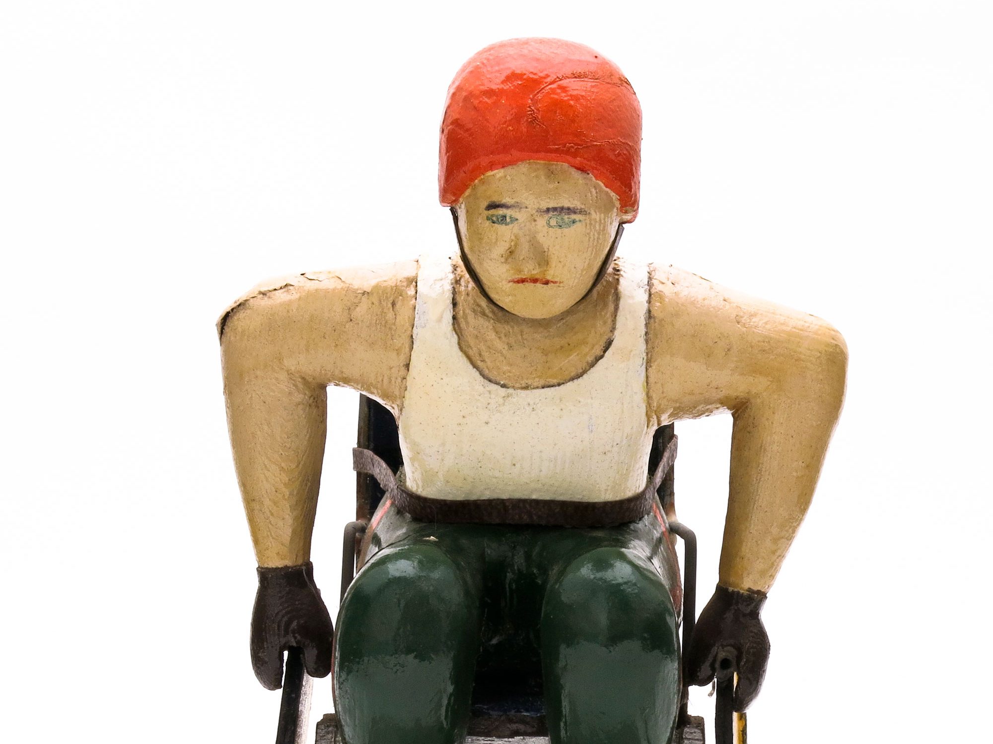Carved painted wooden figure, on a base, of a male athlete in a wheelchair. He is wearing green trousers, a white vest, an orange helmet, and brown gloves. Both his hands are on the wheels of the wheelchair, which has a brown back and solid yellow wheels.