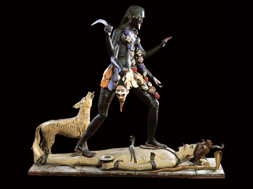 A sculpture of a figure of a woman dancing on a figure of a man who is lying down. The woman has a necklace of heads and is hold a head by its hair in one of her four arms. The man has snakes crawling around his body. There is a tiger behind the woman, that is looking up at her.