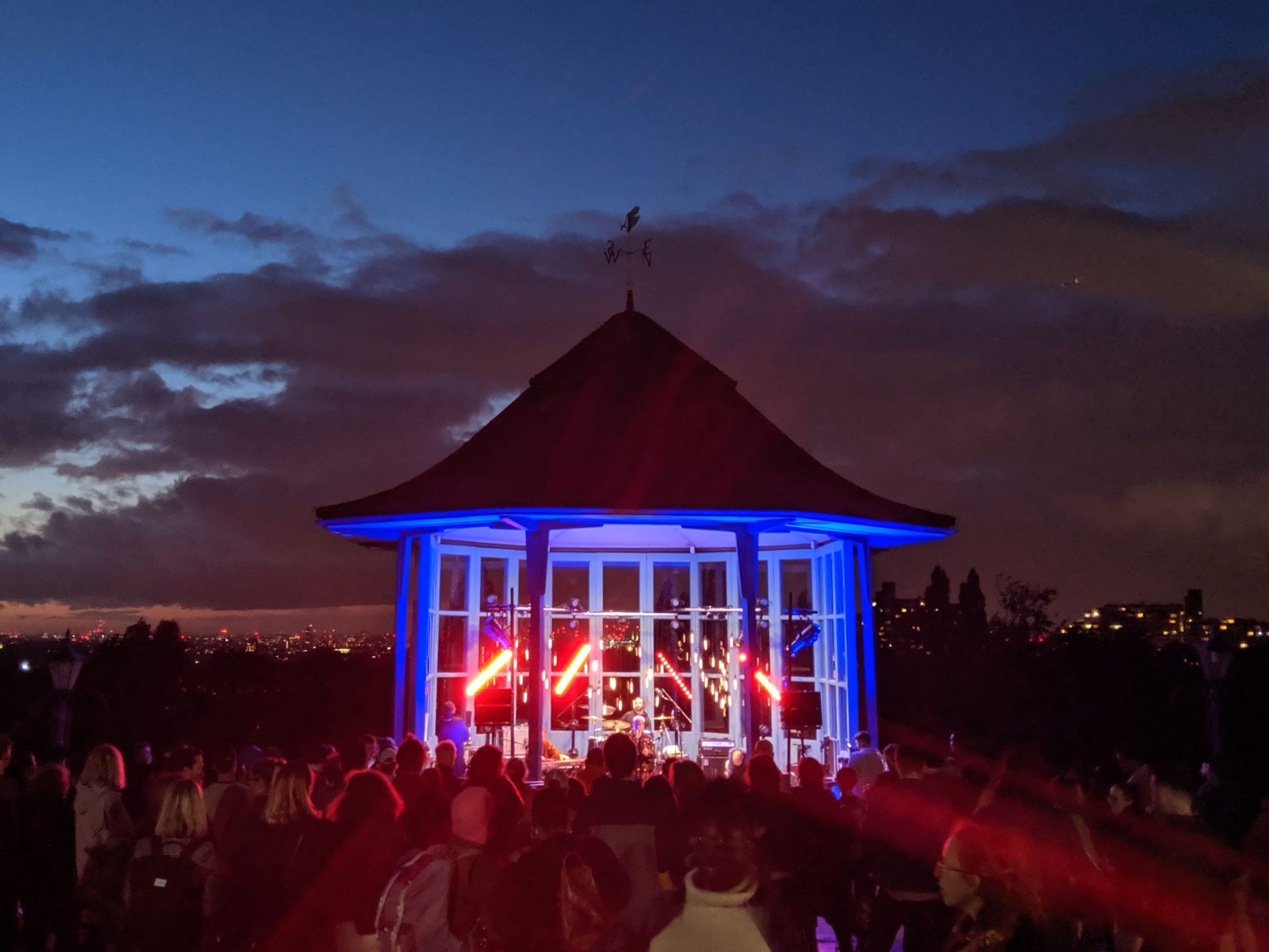 Is is evening and the sky is darkening. There is a small structure with glass windows in front of the camera with red and blue lights shining from it and a performer singing. A crowd is seen looking towards it from behind.