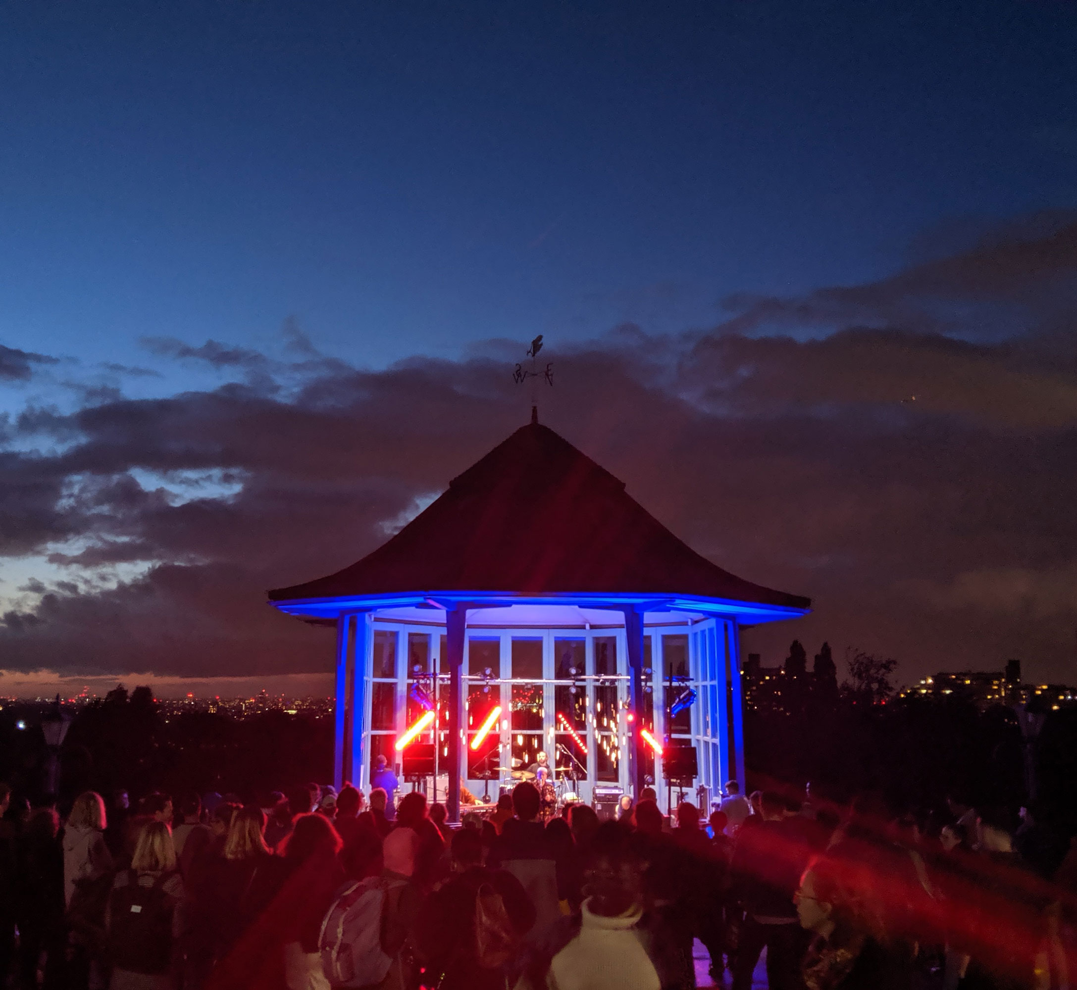 Is is evening and the sky is darkening. There is a small structure with glass windows in front of the camera with red and blue lights shining from it and a performer singing. A crowd is seen looking towards it from behind.