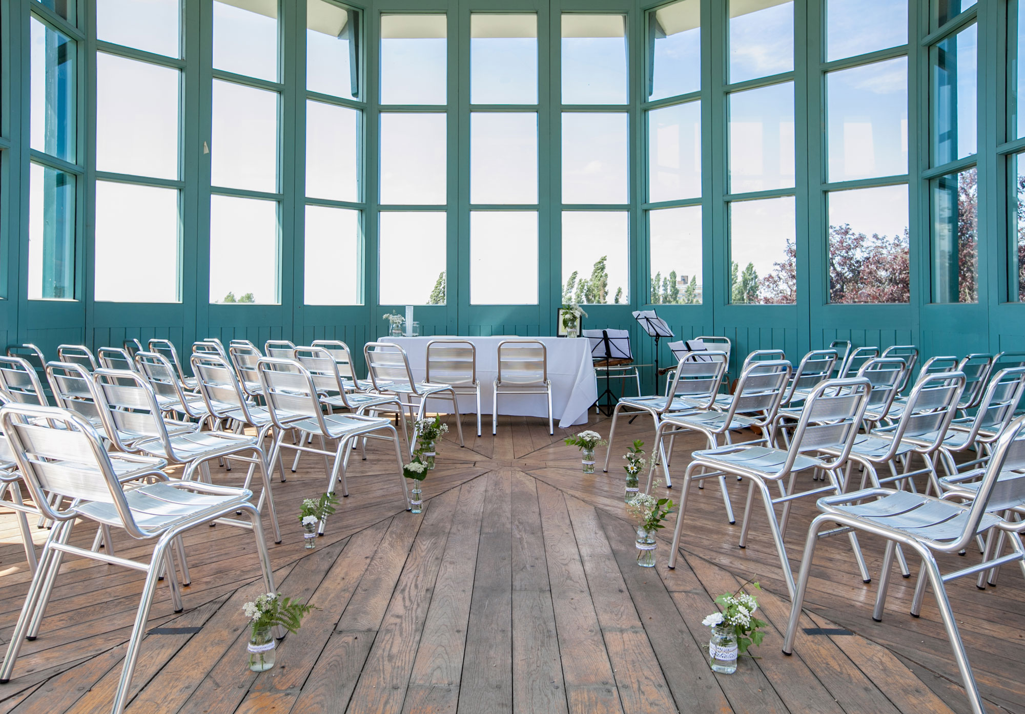 A room surrounded by windows with the surrounds painted blue green. There are chairs facing towards the windows with a table at the end and flowers decorating the wooden floor