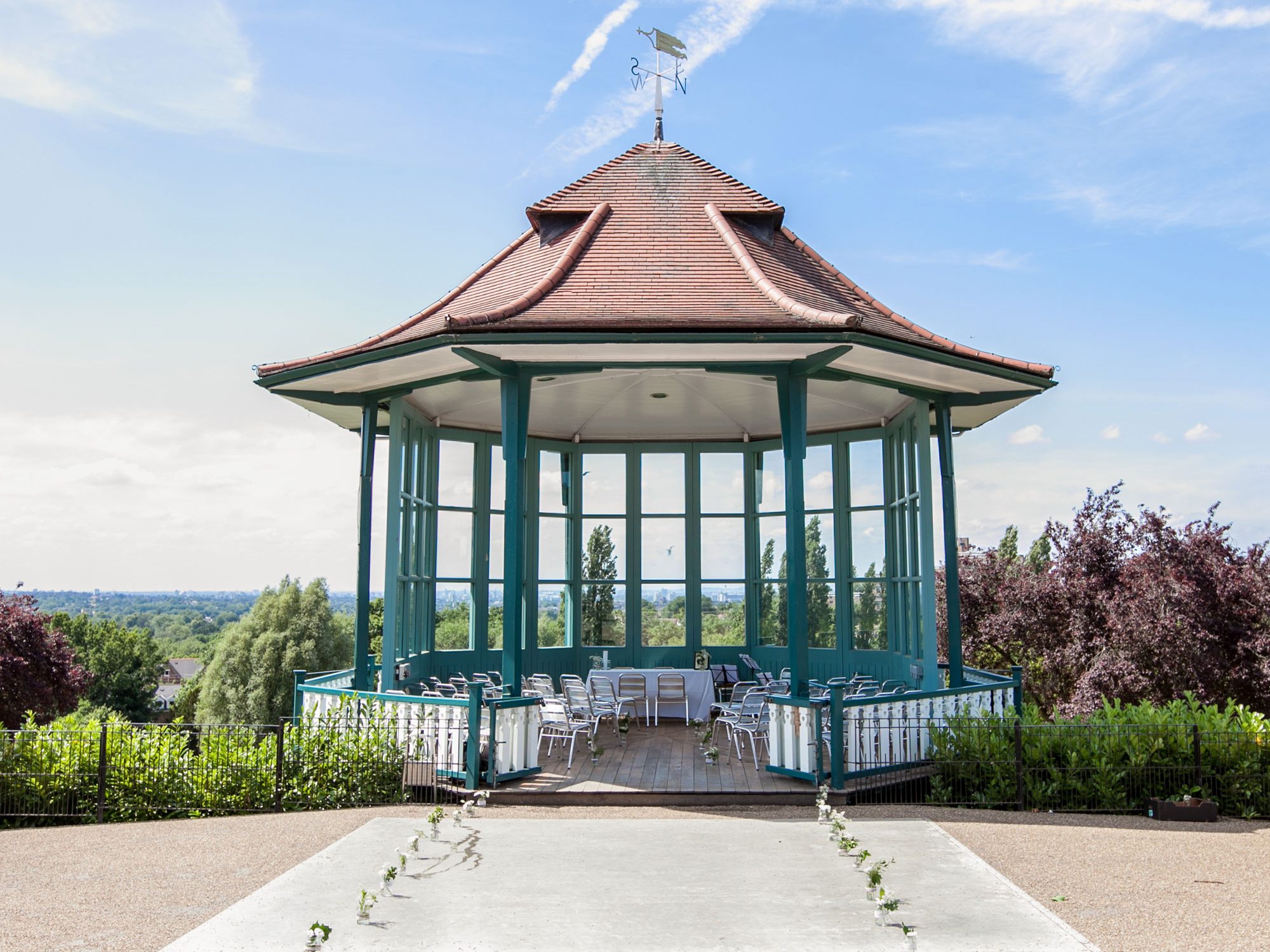 A bandstand, with windows looking out over a view of trees and a city skyline. There are chairs in the bandstand facing the front and jars with flowers in them leading up to the bandstand
