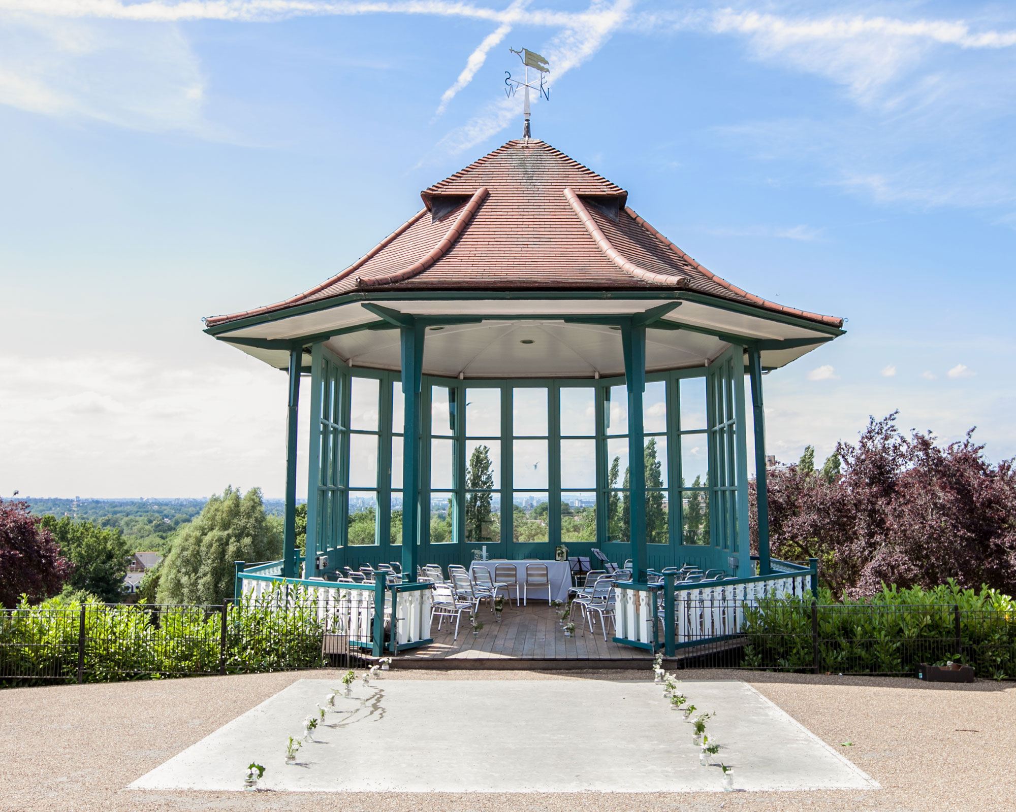 A bandstand, with windows looking out over a view of trees and a city skyline. There are chairs in the bandstand facing the front and jars with flowers in them leading up to the bandstand