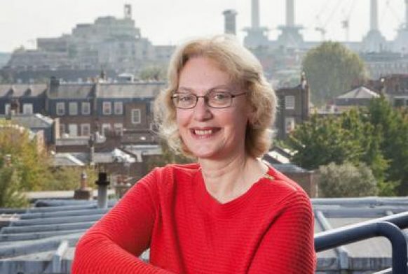 A woman seen from the waist up standing outside with a city skyline in the background. She is wearing a red top, has her arms crossed and is smiling at the camera.