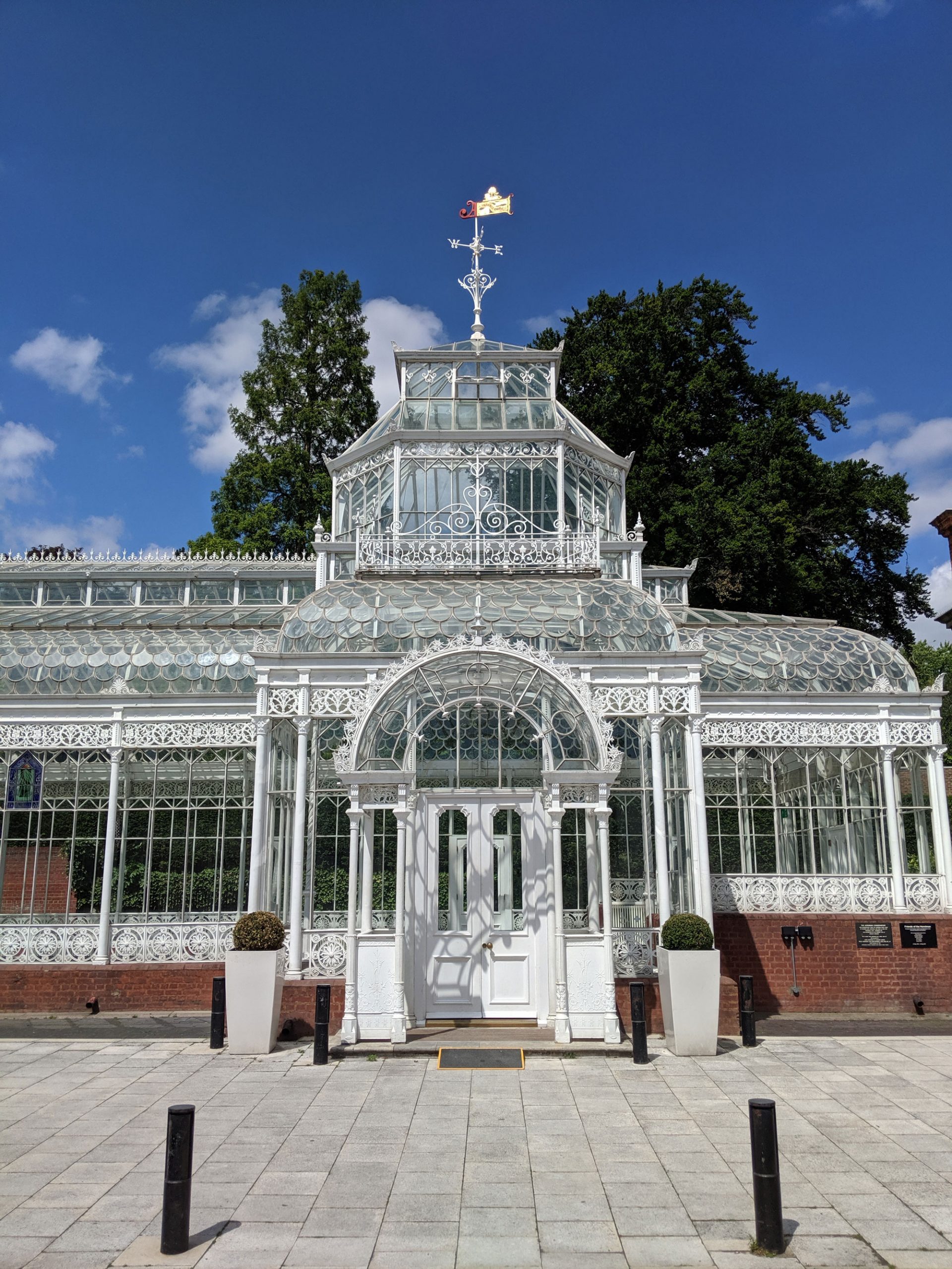 A partial view of an old white metal and glass conservatory seen on a sunny, blue sky day. The conservatory door is in front of us under an arch, with a small glass tower behind it and a weather vane on top. There are two bollards in front of the conservatory.