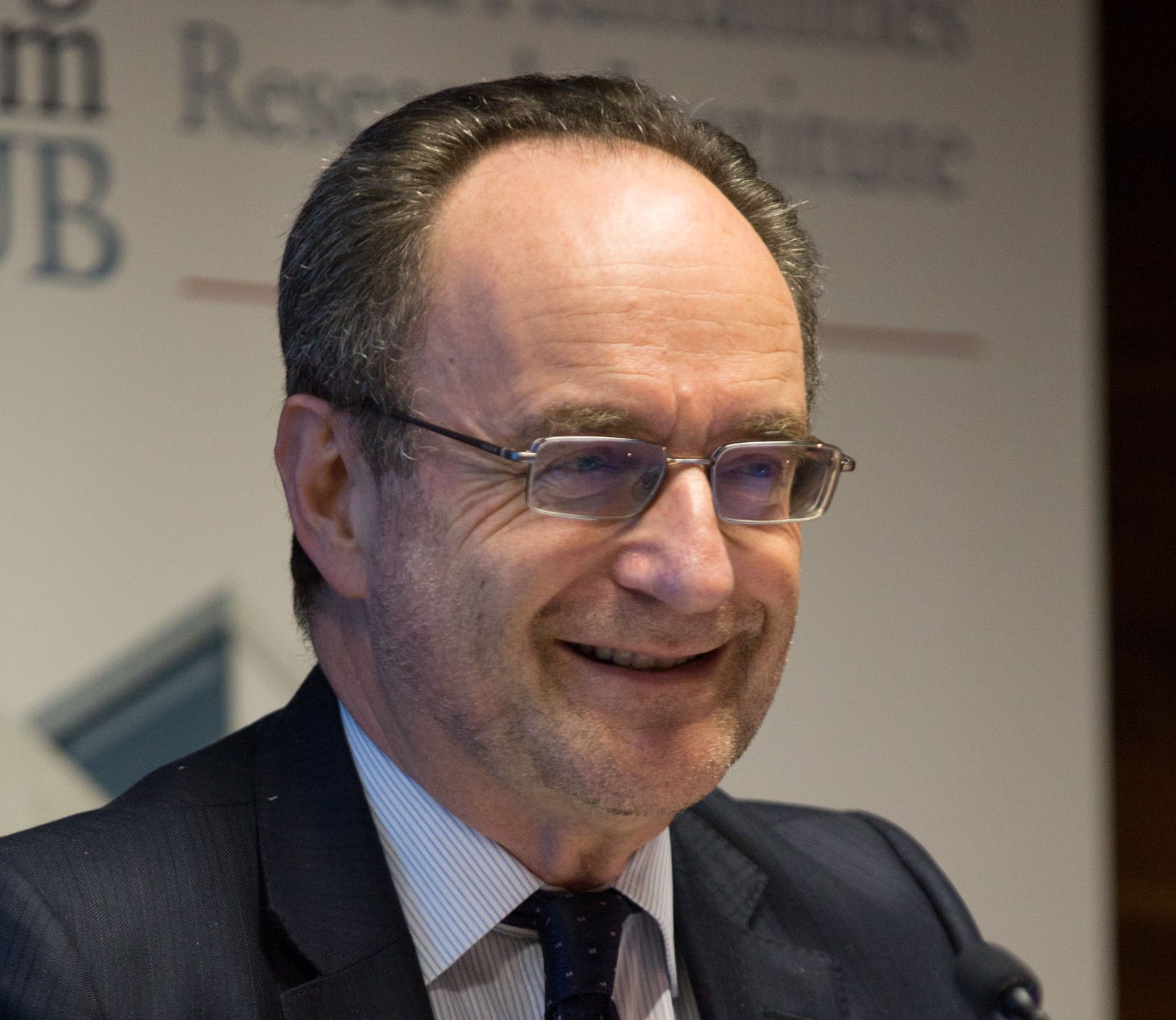 A man seen from the shoulders up smiling, he is stood in front of a board with text written on it (obscured)
