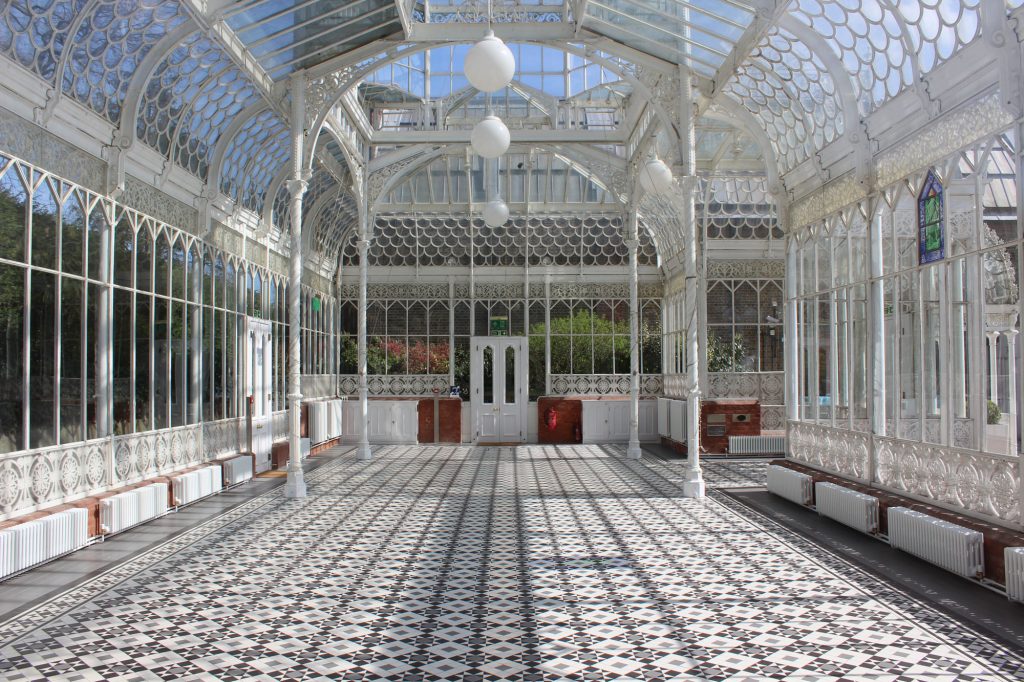 The inside of old old white metal and glass conservatory, with white metal poles around the edges. The floor is a pattern of black and white tiles. There are three globe shaped lamps hanging from the ceiling and there is a double white door at the end. short white radiators edge the space.