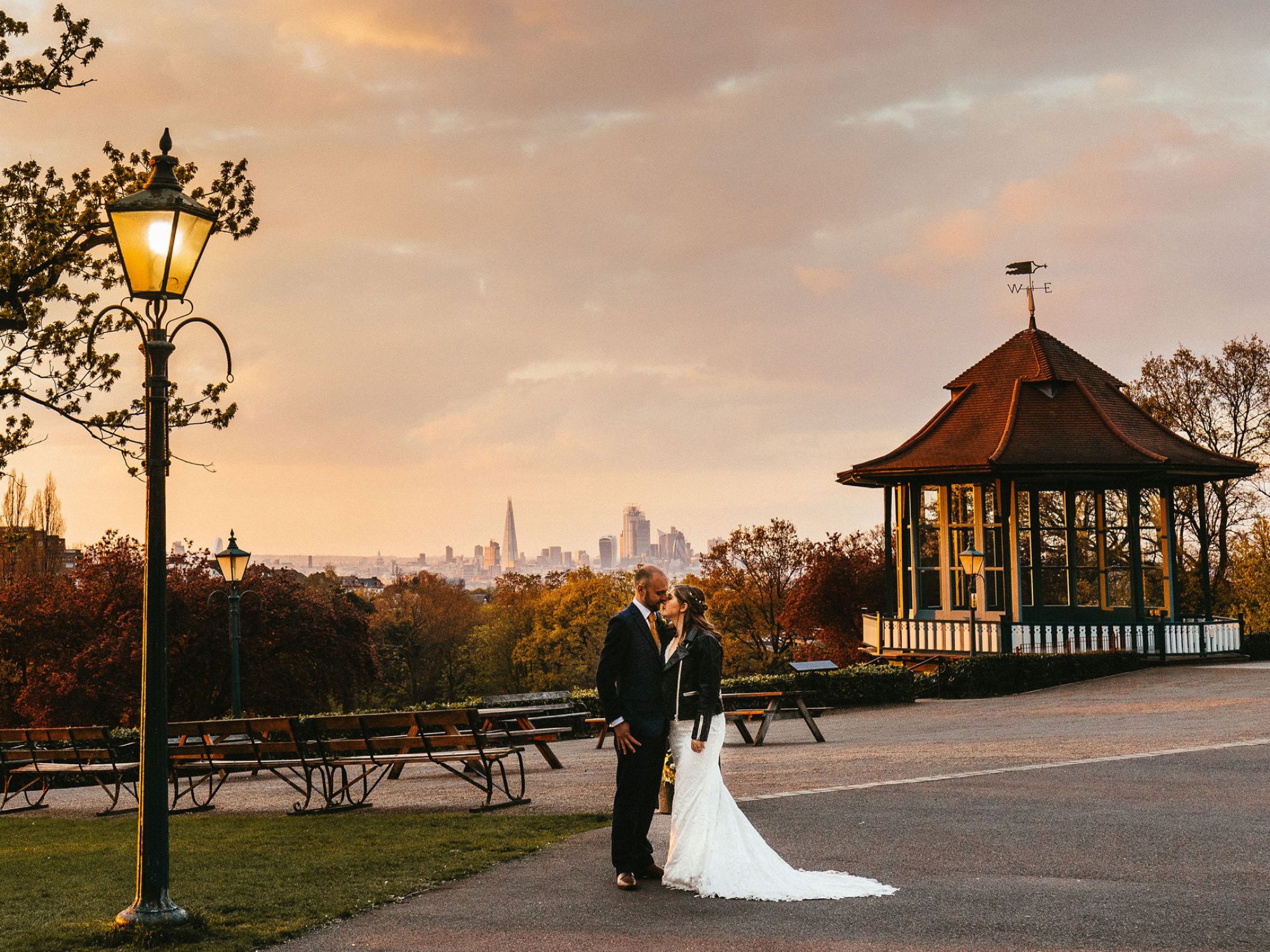 A bride and groom face each other in front of a vista of the London skyline at dusk. There is an old lamppost and tree to the left on a patch of grass, with a terrace stretching out behind them to a bandstand illuminated in the sun