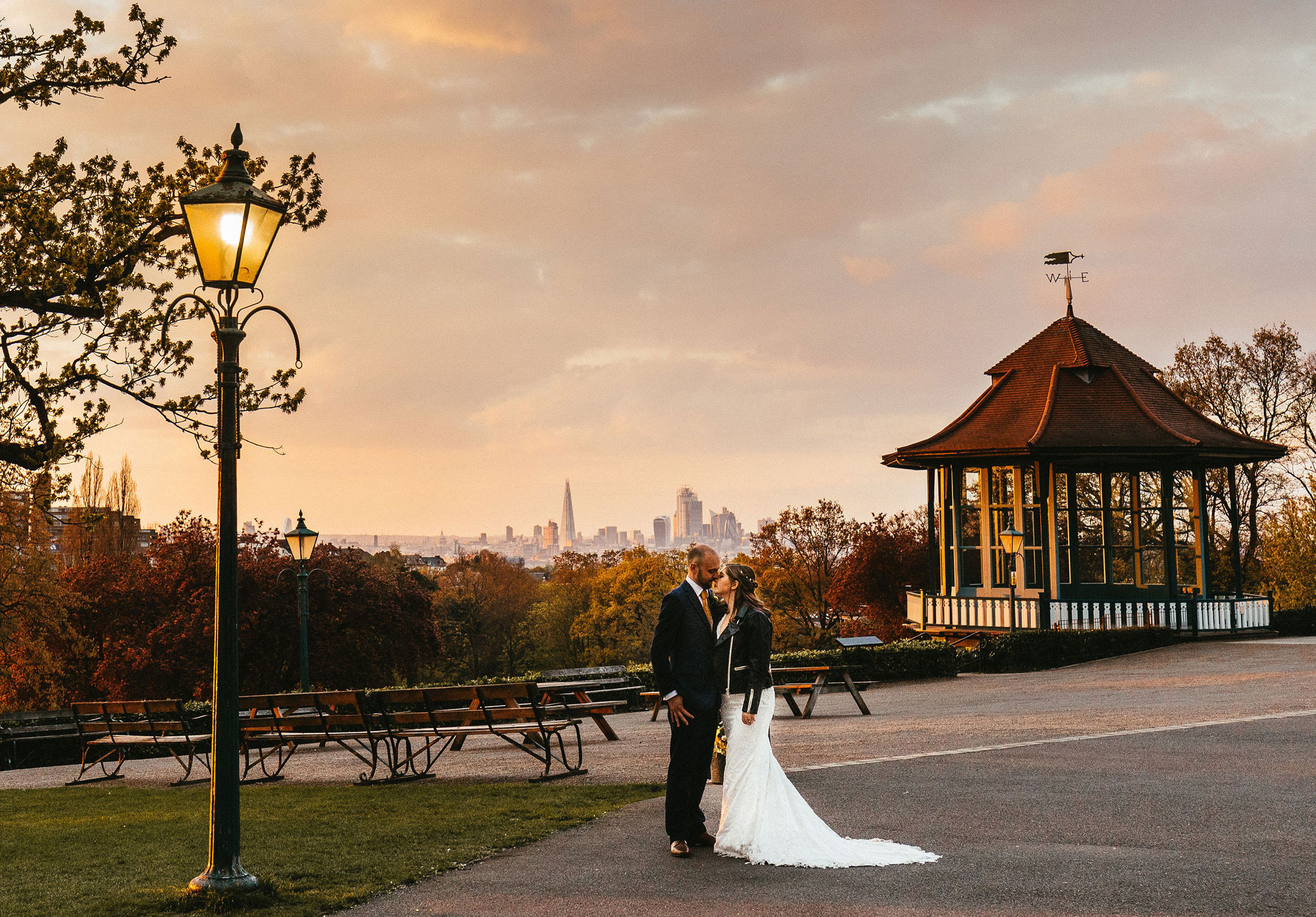 A bride and groom face each other in front of a vista of the London skyline at dusk. There is an old lamppost and tree to the left on a patch of grass, with a terrace stretching out behind them to a bandstand illuminated in the sun