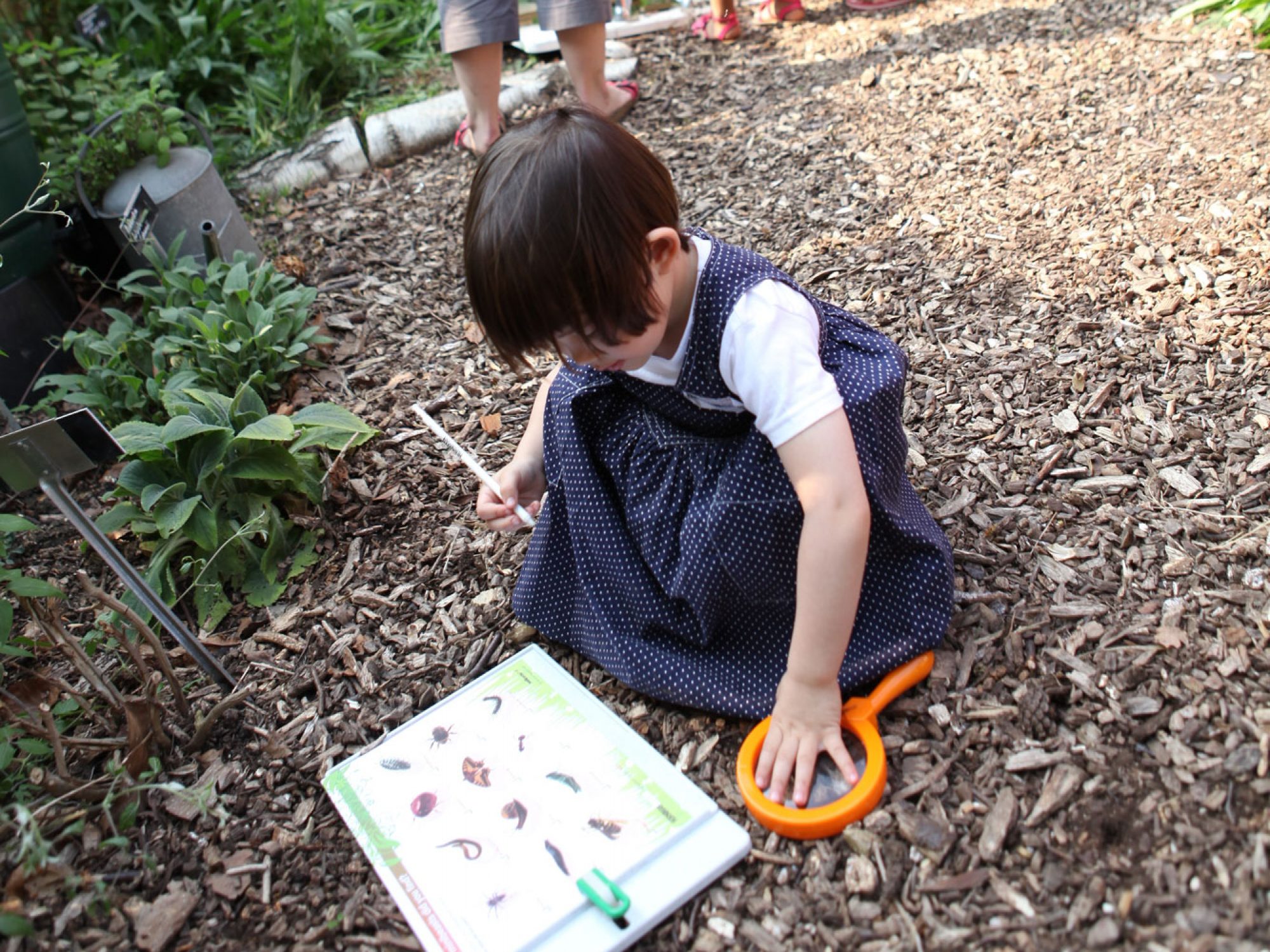 A very young girl crouches down with a pen in her hand, looking at a sheet with pictures on it. Next to her is a magnifying glass. She is on a wood chip strewn trail leading past a fence through trees.