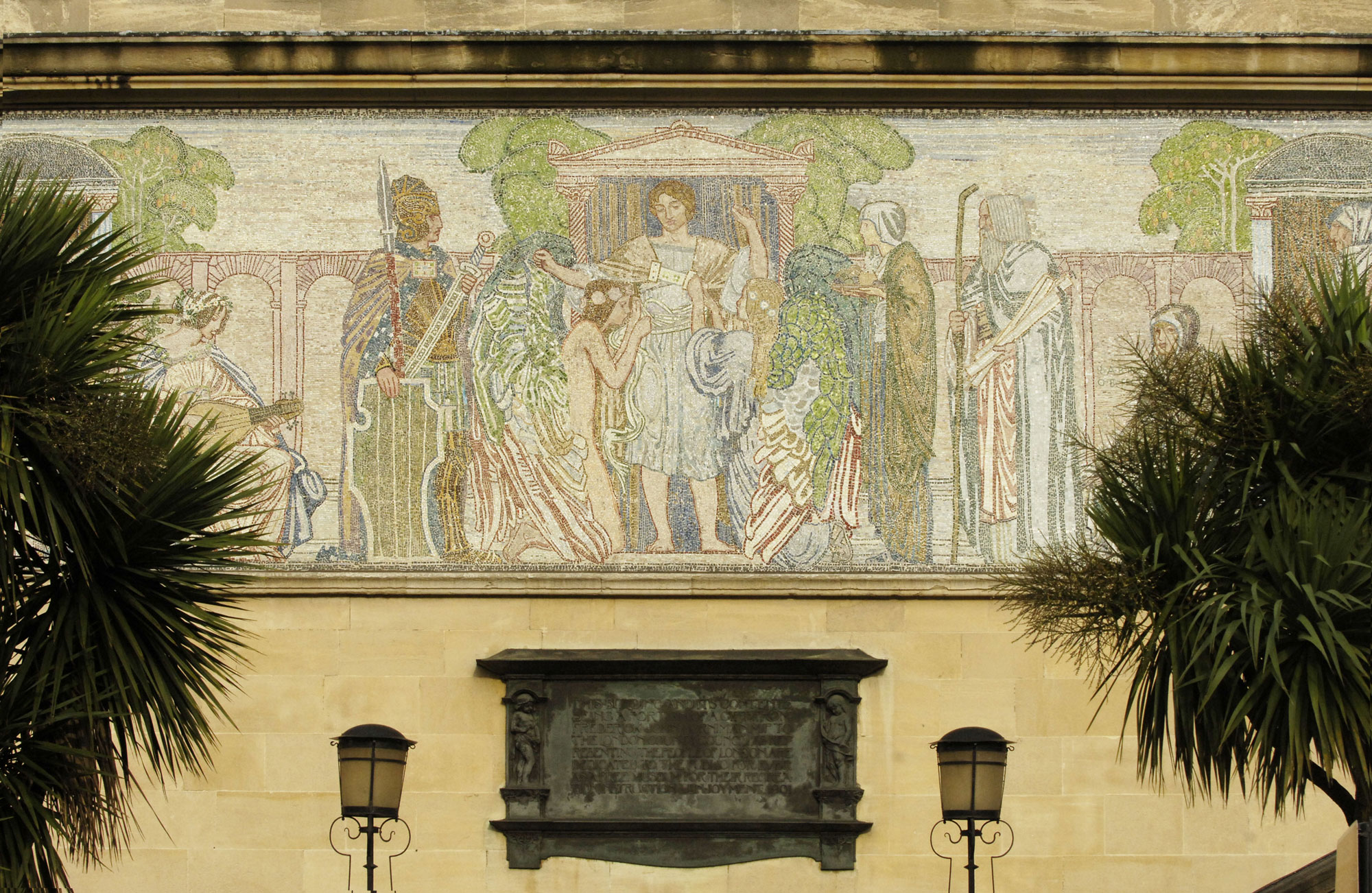 A close up of a mosaic on a cream stone coloured building. The mosaic shows different figures in flowing robes. Beneath it is a plaque and two lamps, and there are palm trees just seen to either side