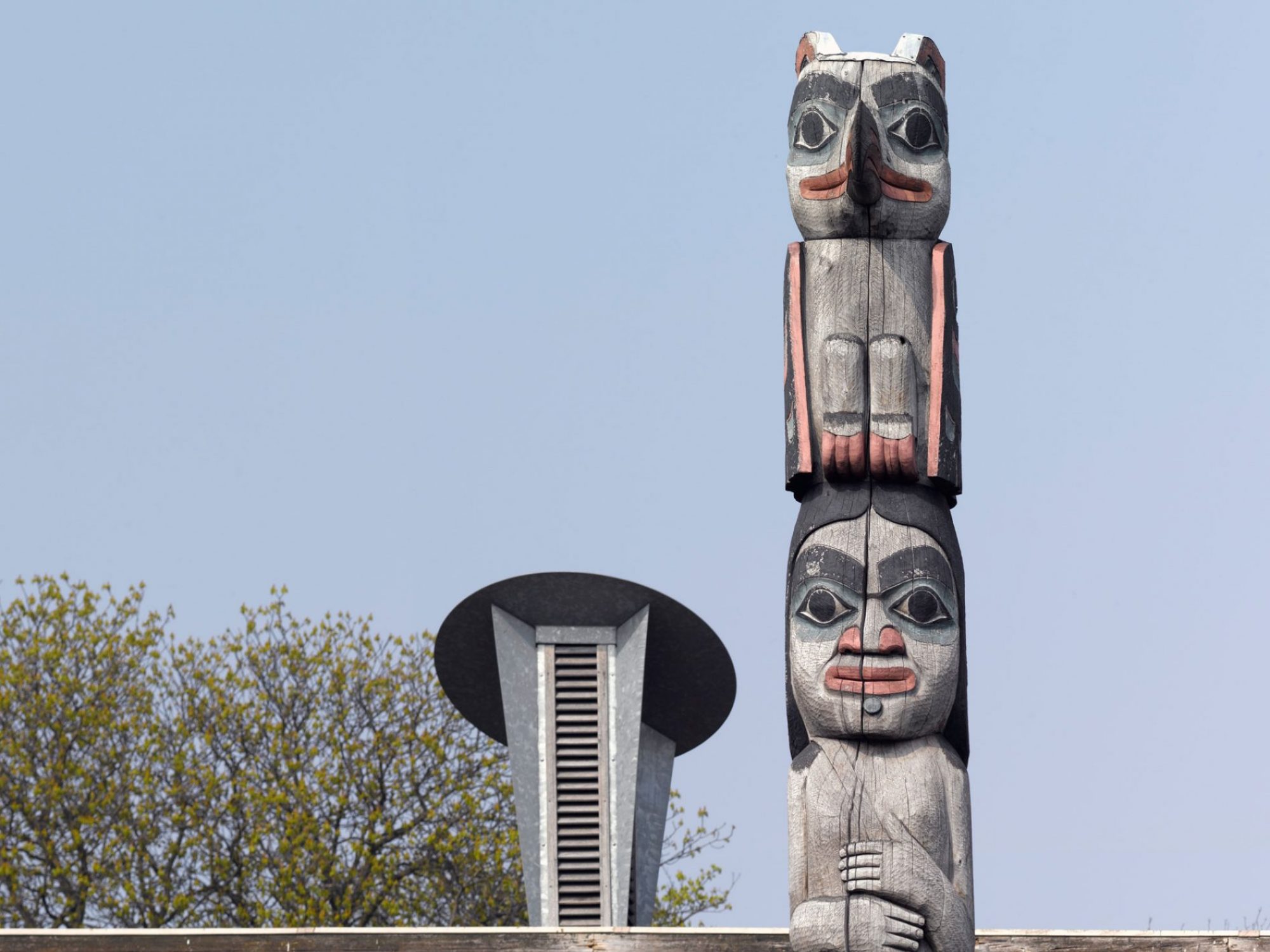 A totem pole made up of three figures. The first is a bear, the second is a woman and the third is a bird. Behind the pole is a building with grass growing on the roof and tall air vents.
