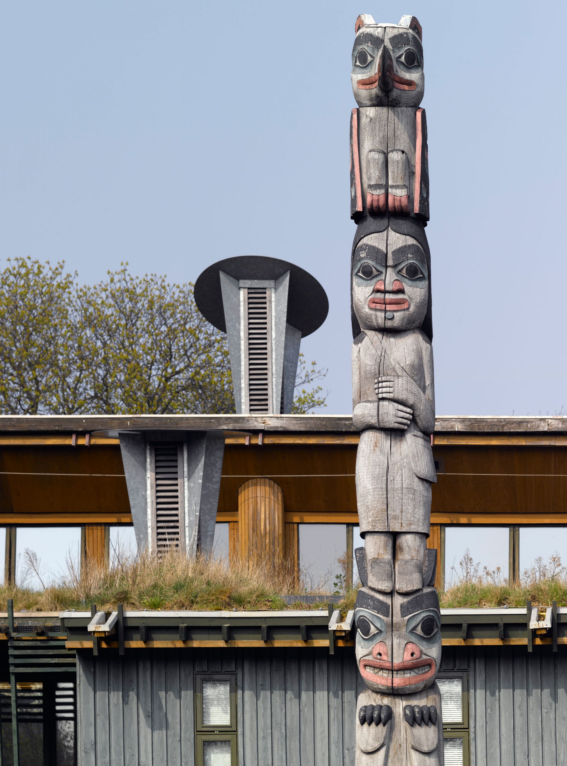 A totem pole made up of three figures. The first is a bear, the second is a woman and the third is a bird. Behind the pole is a building with grass growing on the roof and tall air vents.