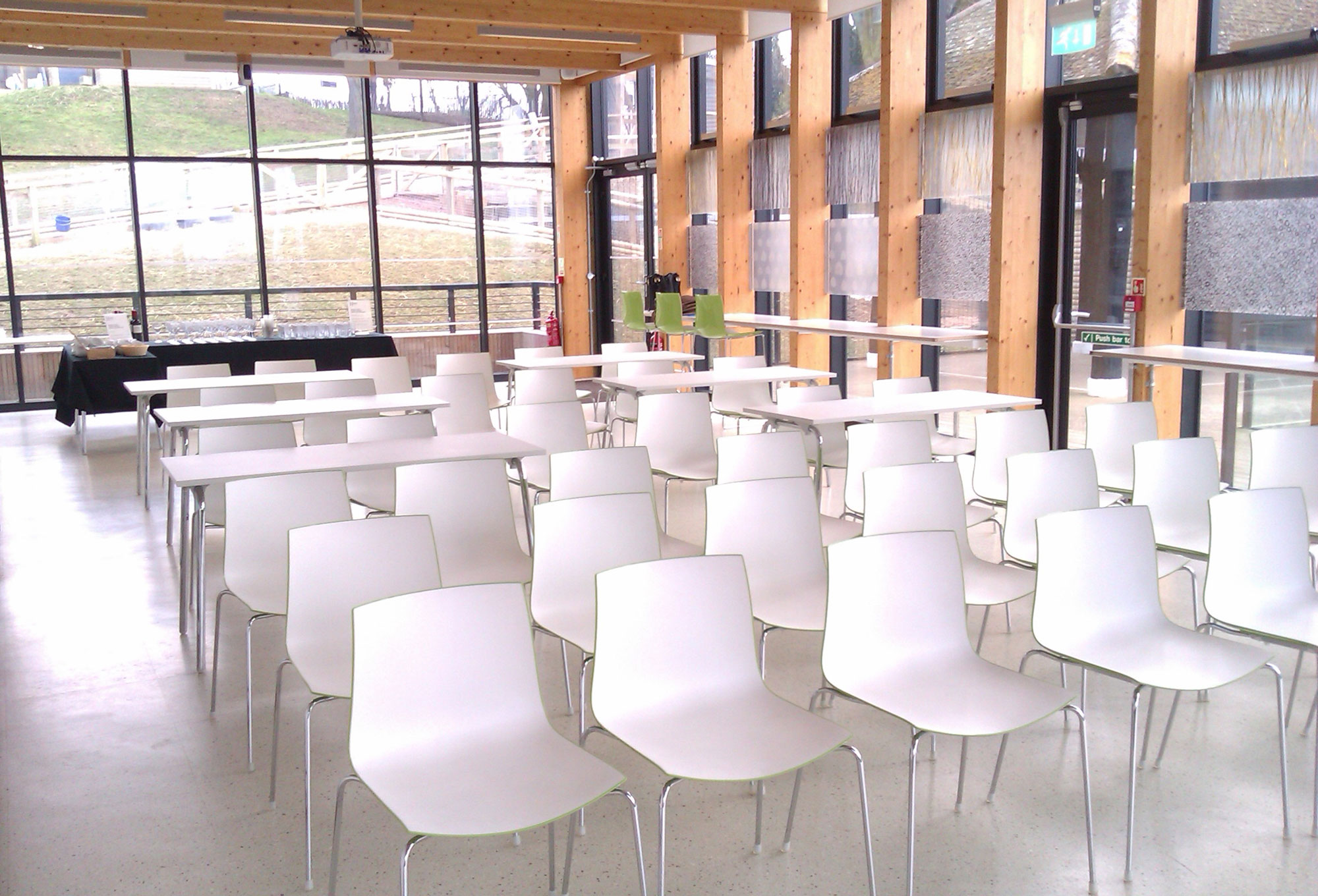 A light room is full of white chairs all facing towards the camera. The room has floor to ceiling glass windows at the back and side that is visible, with some greenery just seen outside