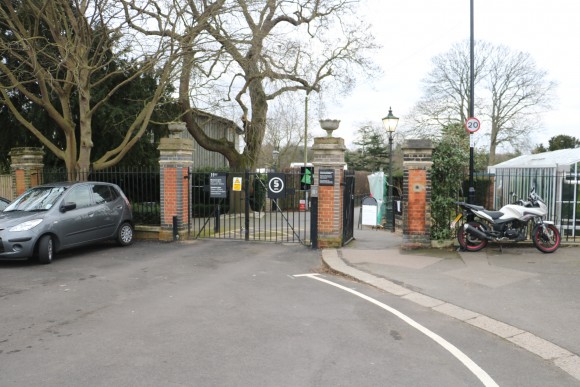 An old fashioned looking metal gate with brick supports to either side. There is a grey car parked next to them on the left and a motorbike parked to the right. A pedestian gate is open to the right hand side. The gates themselves are closed and a sign saying 5 miles per hour is just visible.