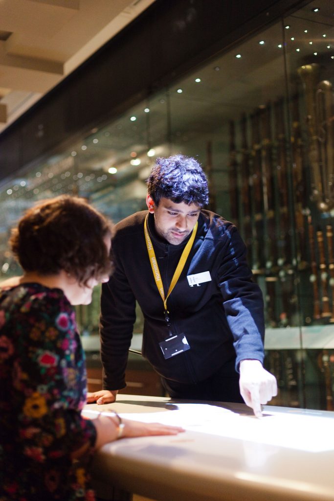 A visitor host in the Horniman music gallery is helping a woman seated at a table.