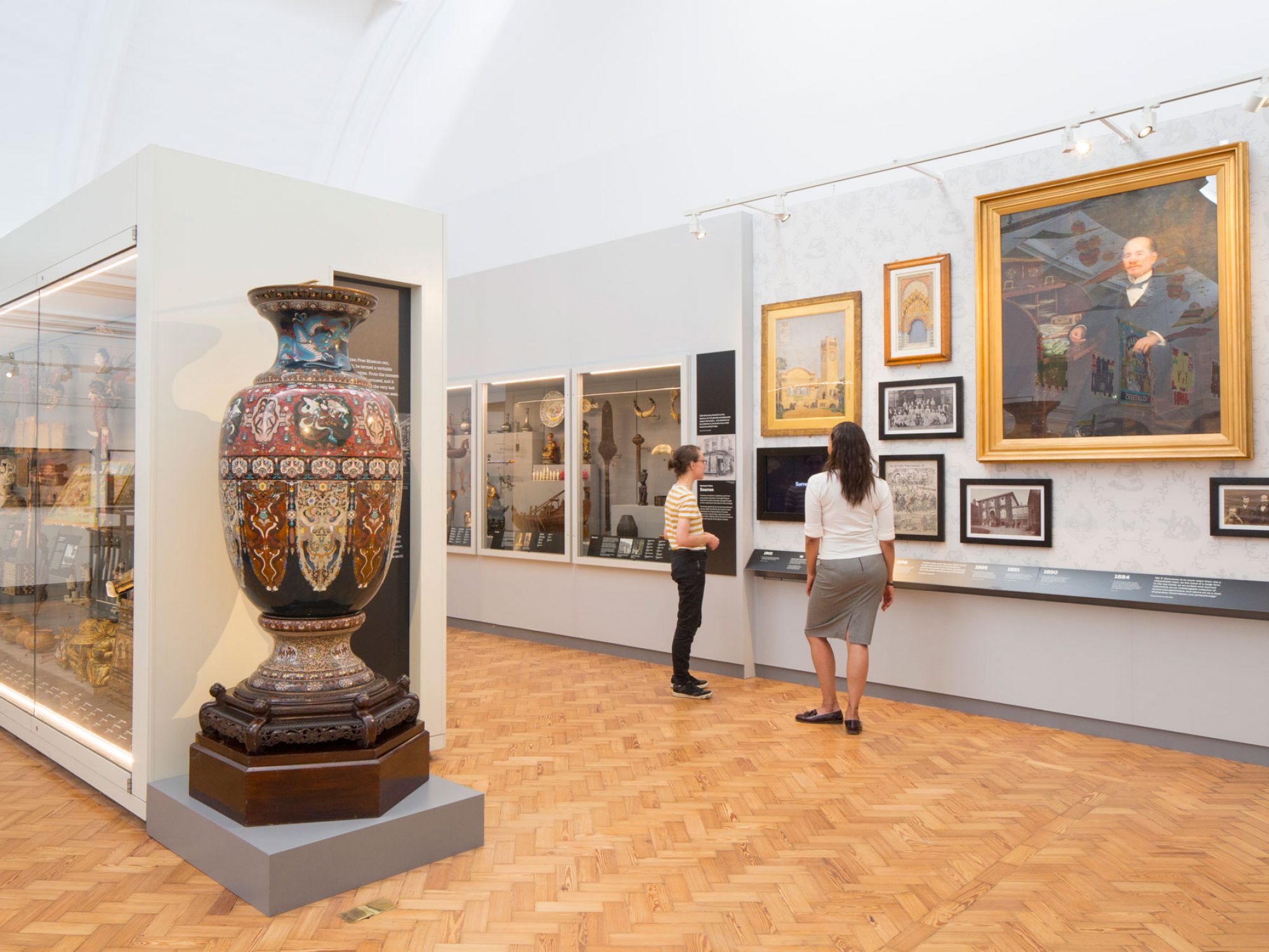 A wide gallery space with pictures on the walls and cabinets with many objects in them. A large vase taller than a person is to the front of the camera and two woman are looking at paintings