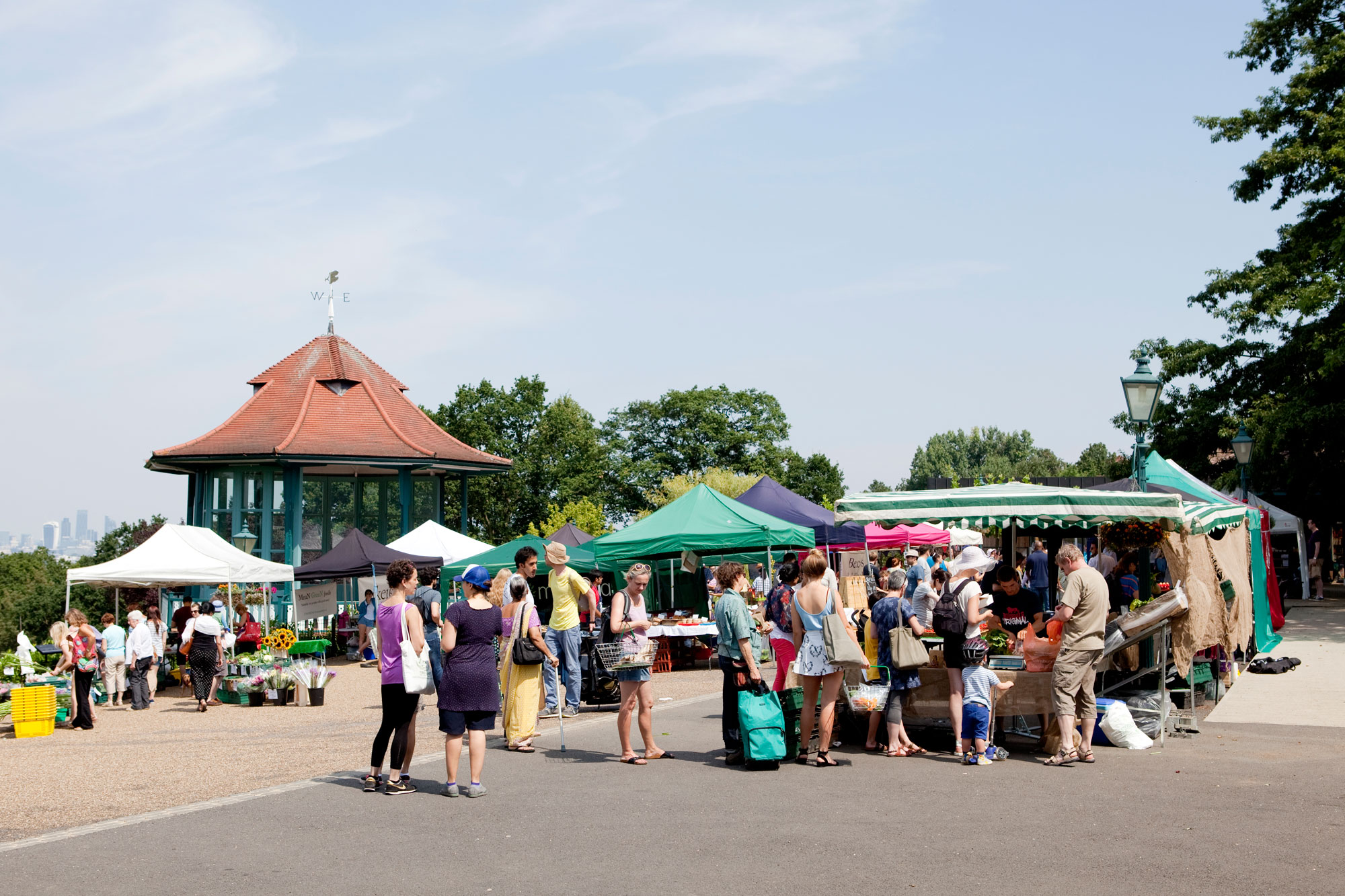 A group of stalls in a terrace outdoors next to a bandstand. There are people queuing at the stalls and the day is sunny and blue