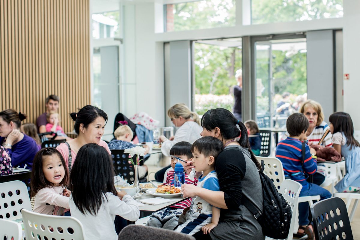A busy cafe with table and chairs full of adults and children eating. The space is light and airy with glass walls.