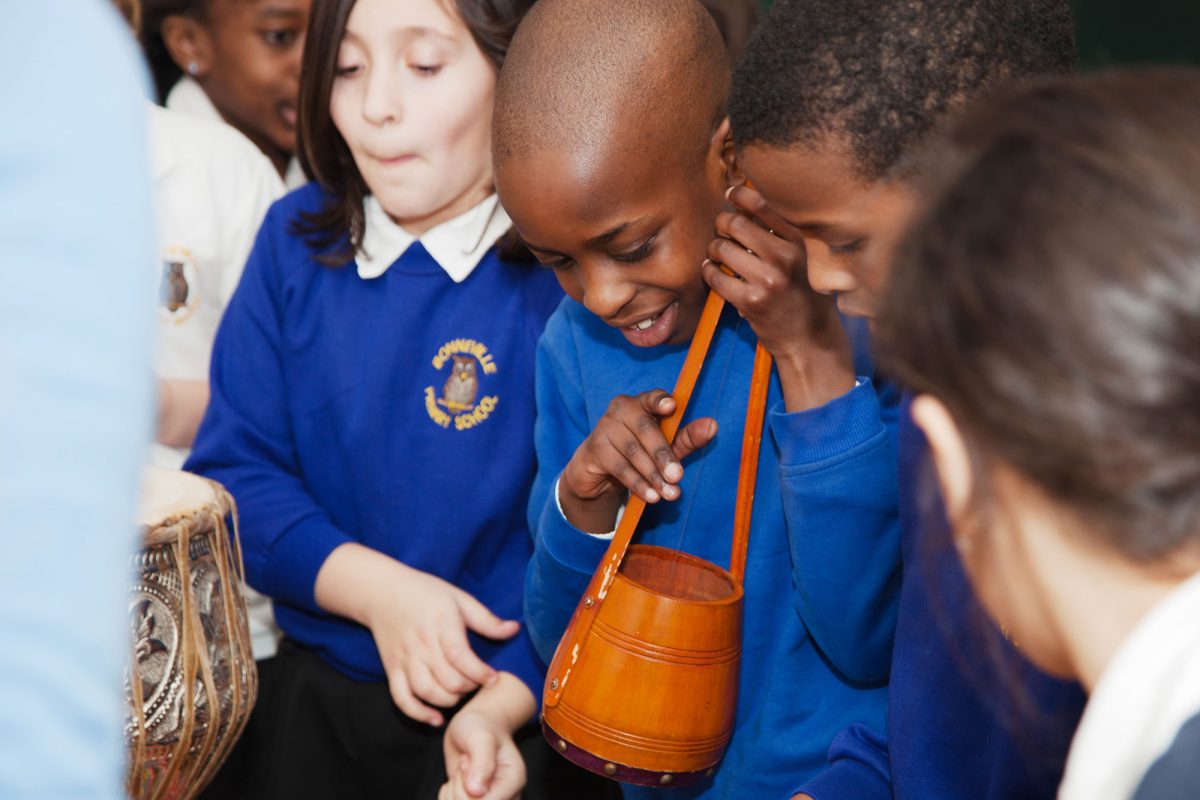 Four children are grouped around some instruments wearing blue jumpers. One child is holding a stringed drum shaped instrument and is smiling. There is a drum just seen in front of them.