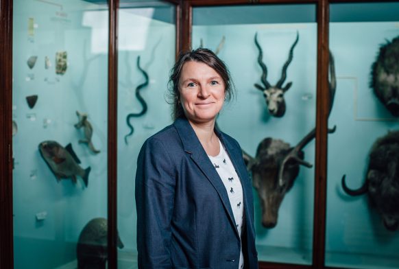 A woman seen from the waist up is wearing a blue jacket and is smiling. She is stood in front of some blue museum cases with taxidermy animals in them like fish, deer and antelope