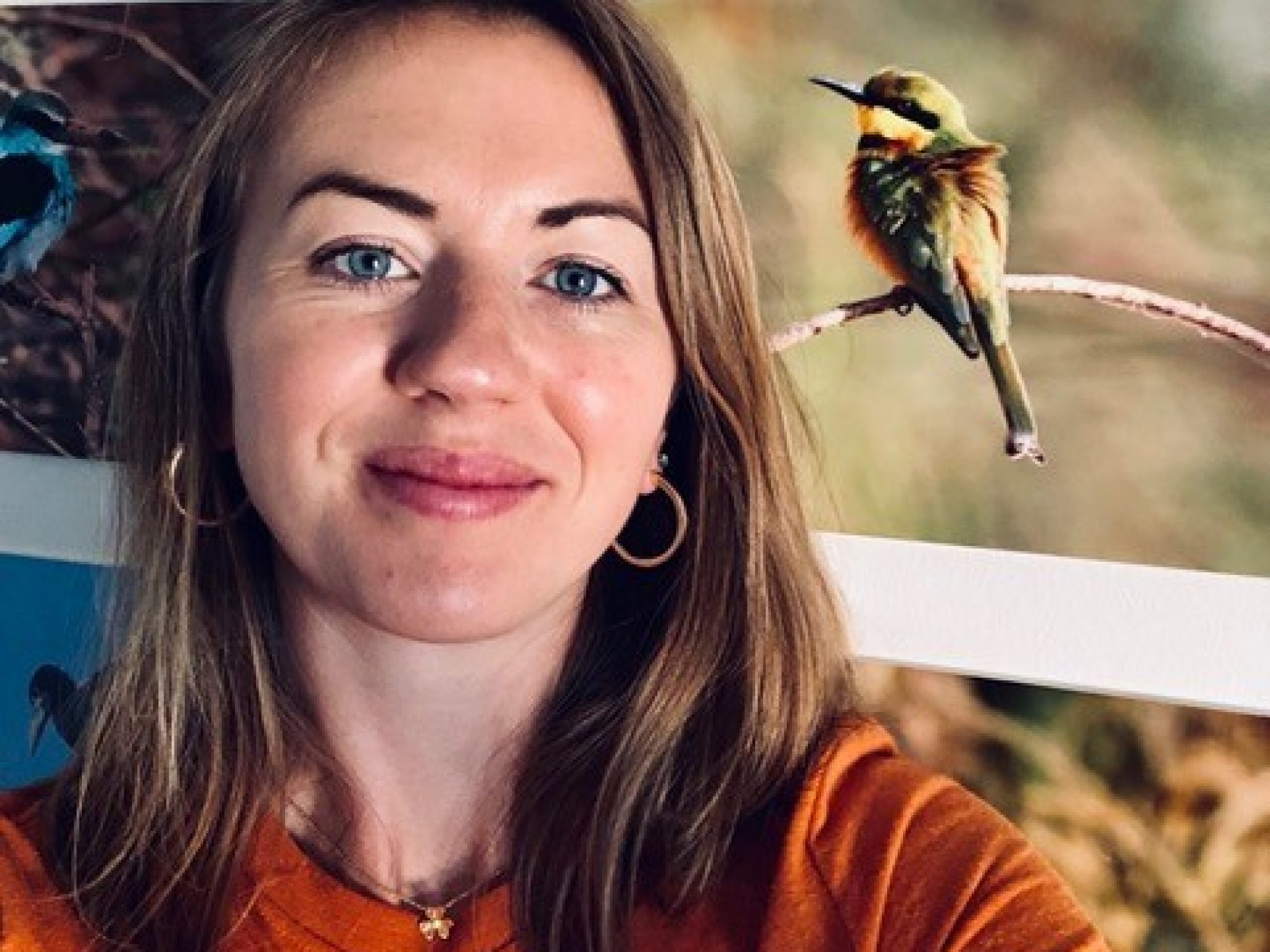A woman smiles at the camera, taking a selfie. She is in front of pictures of nature, like plants and birds