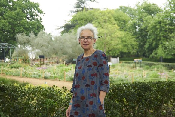 A woman stands in front of a low hedge, with a garden behind it. She is wearing a blue dress, and has white hair and glasses