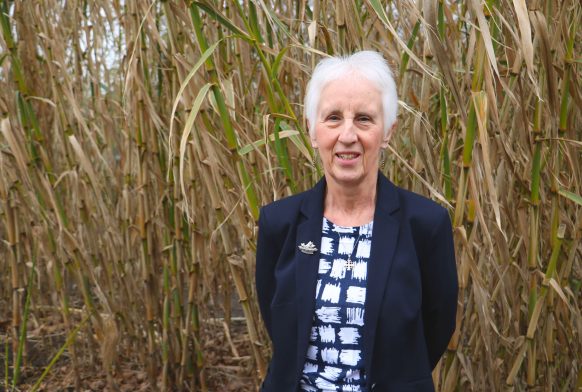 A woman is seen from the waist up wearing a dark blue jacket. She is smiling and stood in front of some bamboo outside.