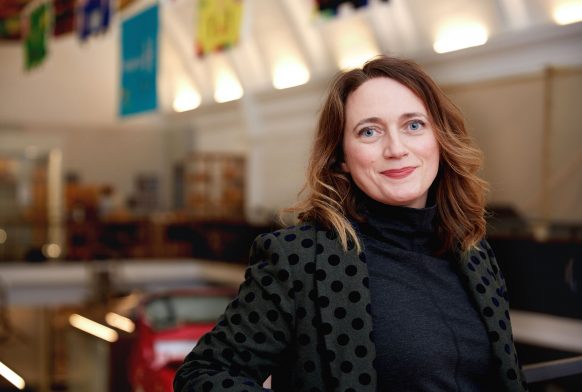 A woman is seen from mid torso up. She is wearing a black spotted jacket and is smiling. Behind her is a gallery which is blurred.