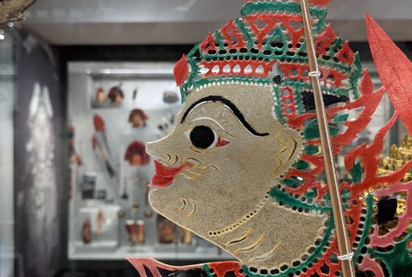 A shadow puppet made of very thin paper, as seen through a glass case. the puppet is wearing an ornate red outfit and we see only the face but it is hard to tell whether they are male or female