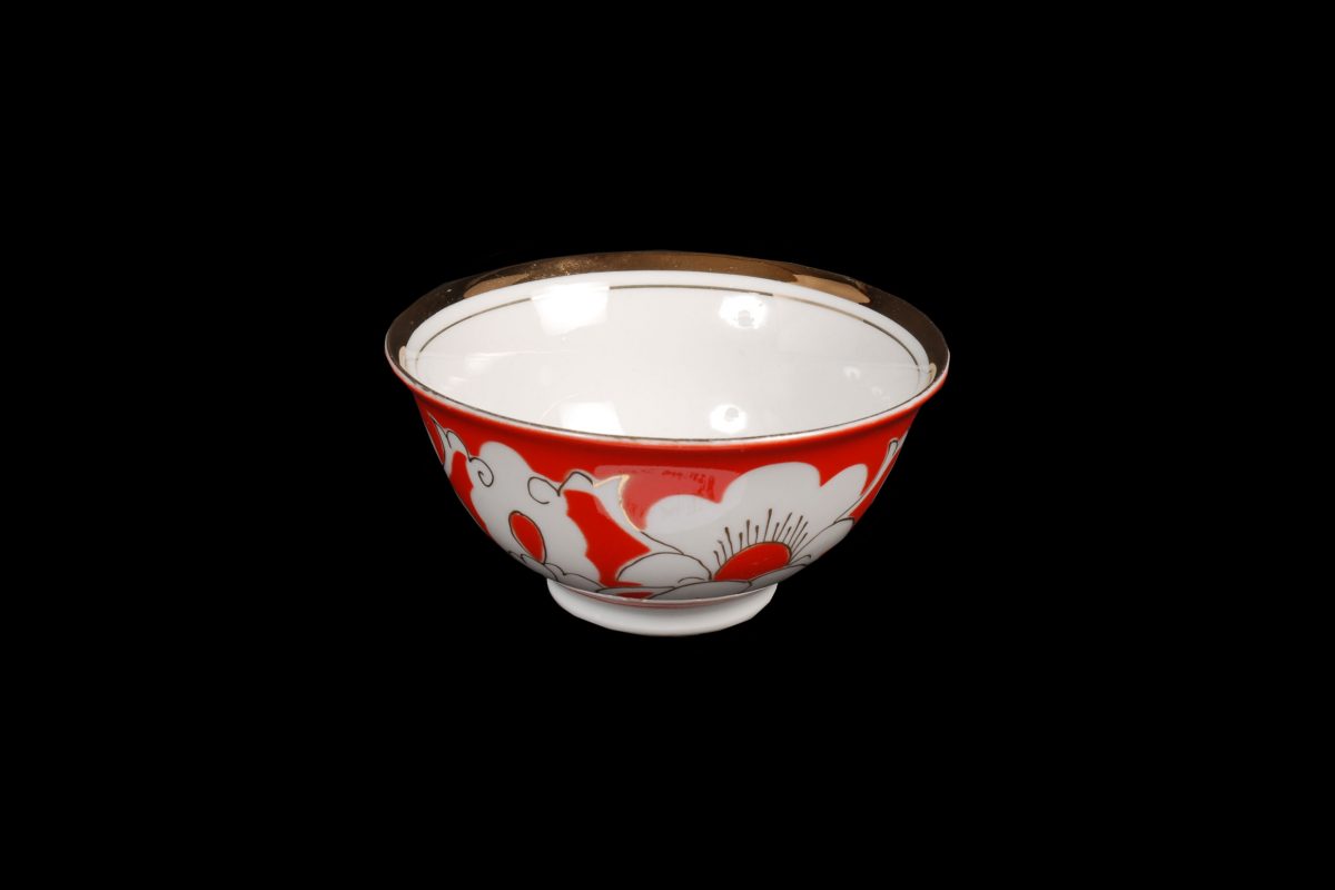 Small bowl for tea or spirits. Part of a factory made pottery tea set.Typical decoration with white motifs (cotton plants) on an orange-red ground, trimmed with a golden band around the rim.