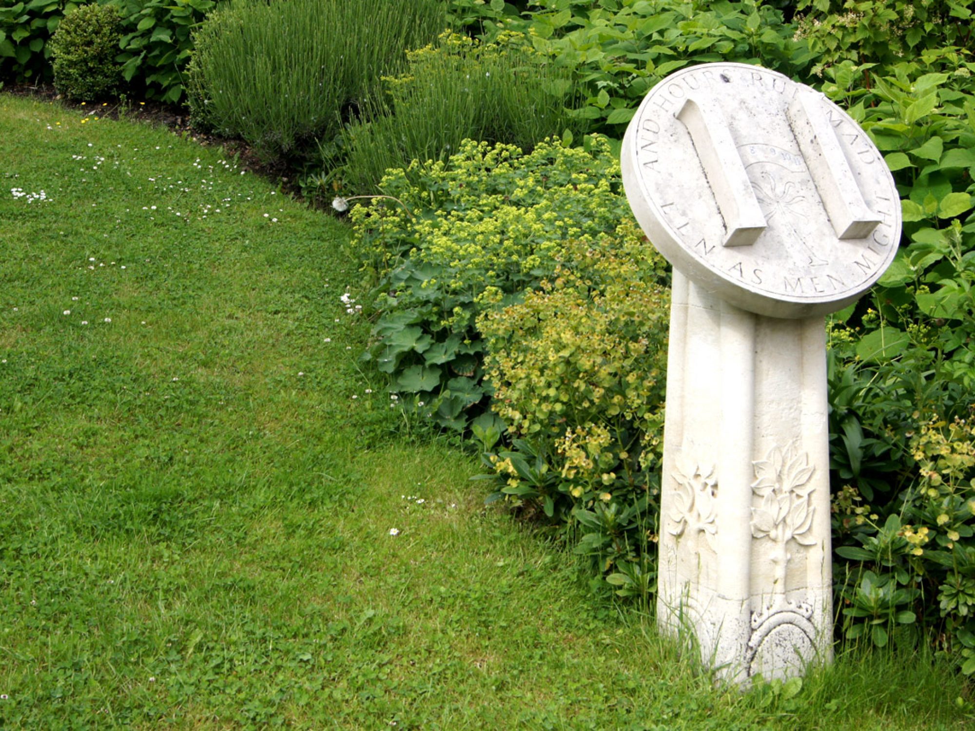 A stone sundial in a garden with a H on the front