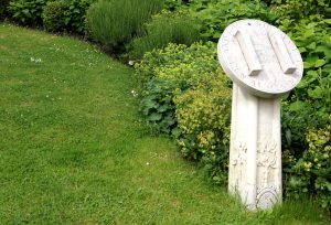 A stone sundial in a garden with a H on the front