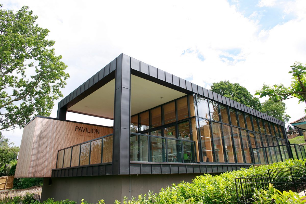 Glass rectangle building on hill surrounded by trees with a cloudy sky behind.