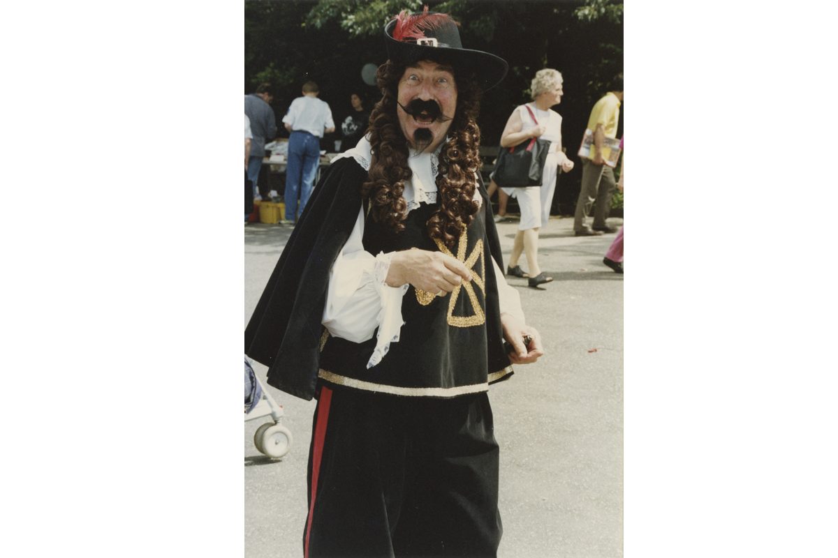 Man in costume with long hear, and mostache. People are walking behind him and shopping at market stall.