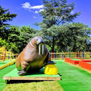A model of a walrus standing on a yellow golf ball is in a garden golf course on a sunny day