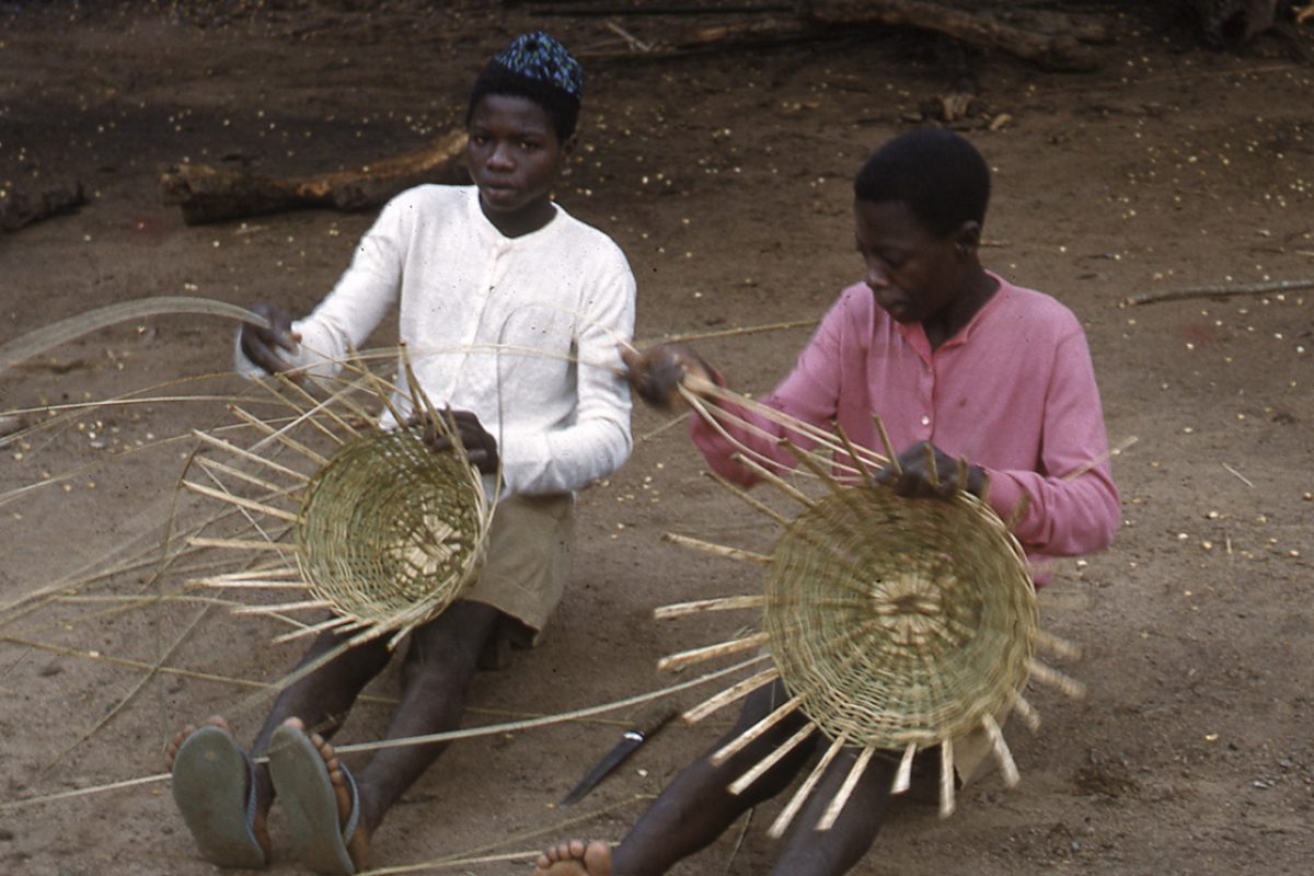 Two women sitting on bare ground weaving baskets on their laps.