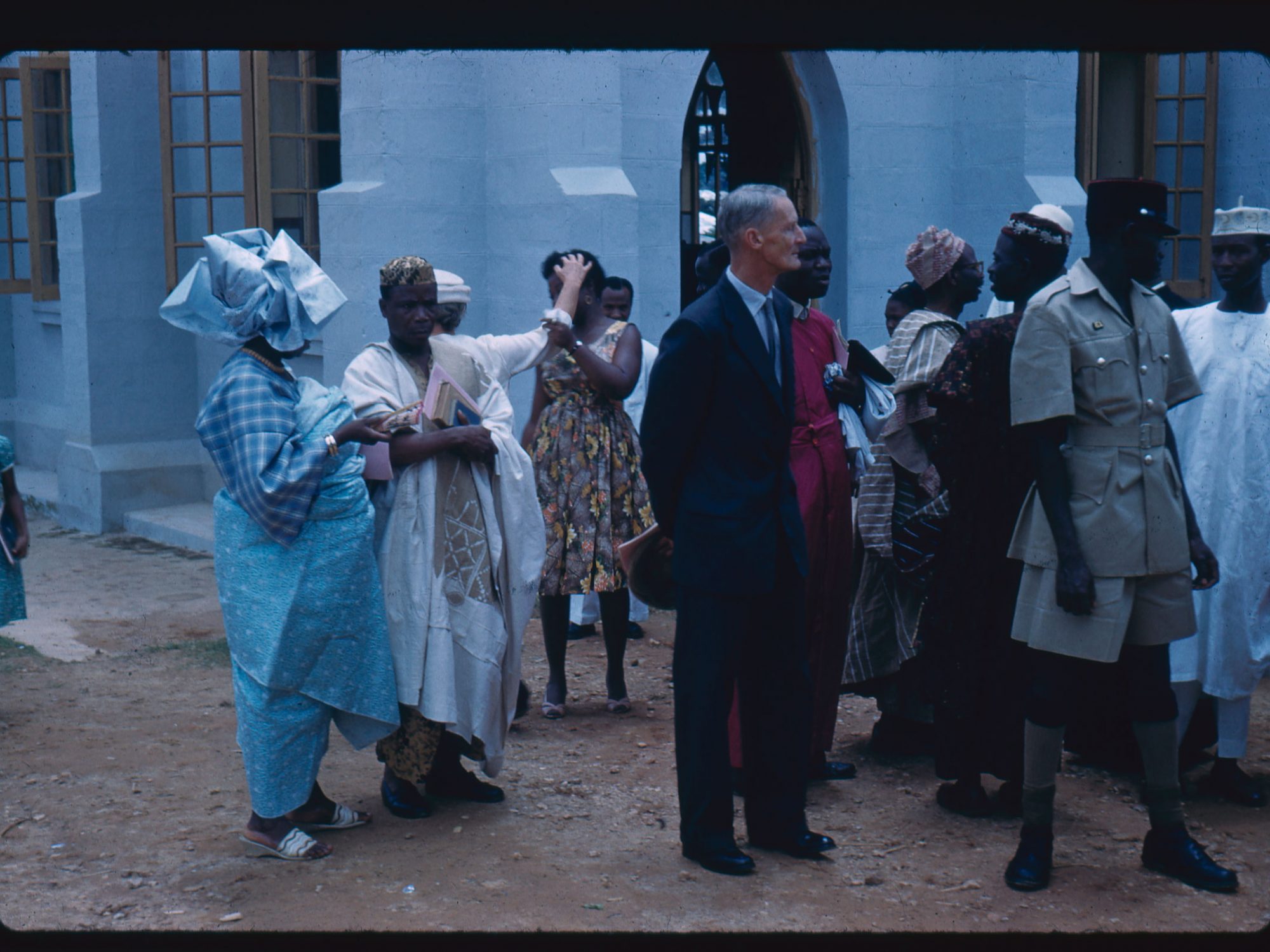 A old colour photo of a formal or celebratory occasion. Two people to the left are in blue and white clothes. A man to the centre is in a suit and to his right is a man in a khaki uniform. Behind him are a group of people wearing robes. Behind the people in the foreground is a woman in a sun dress who is holding a second woman's arm, who is in a white dress. The woman in the white dress is reaching for her and looking away from her at the same time